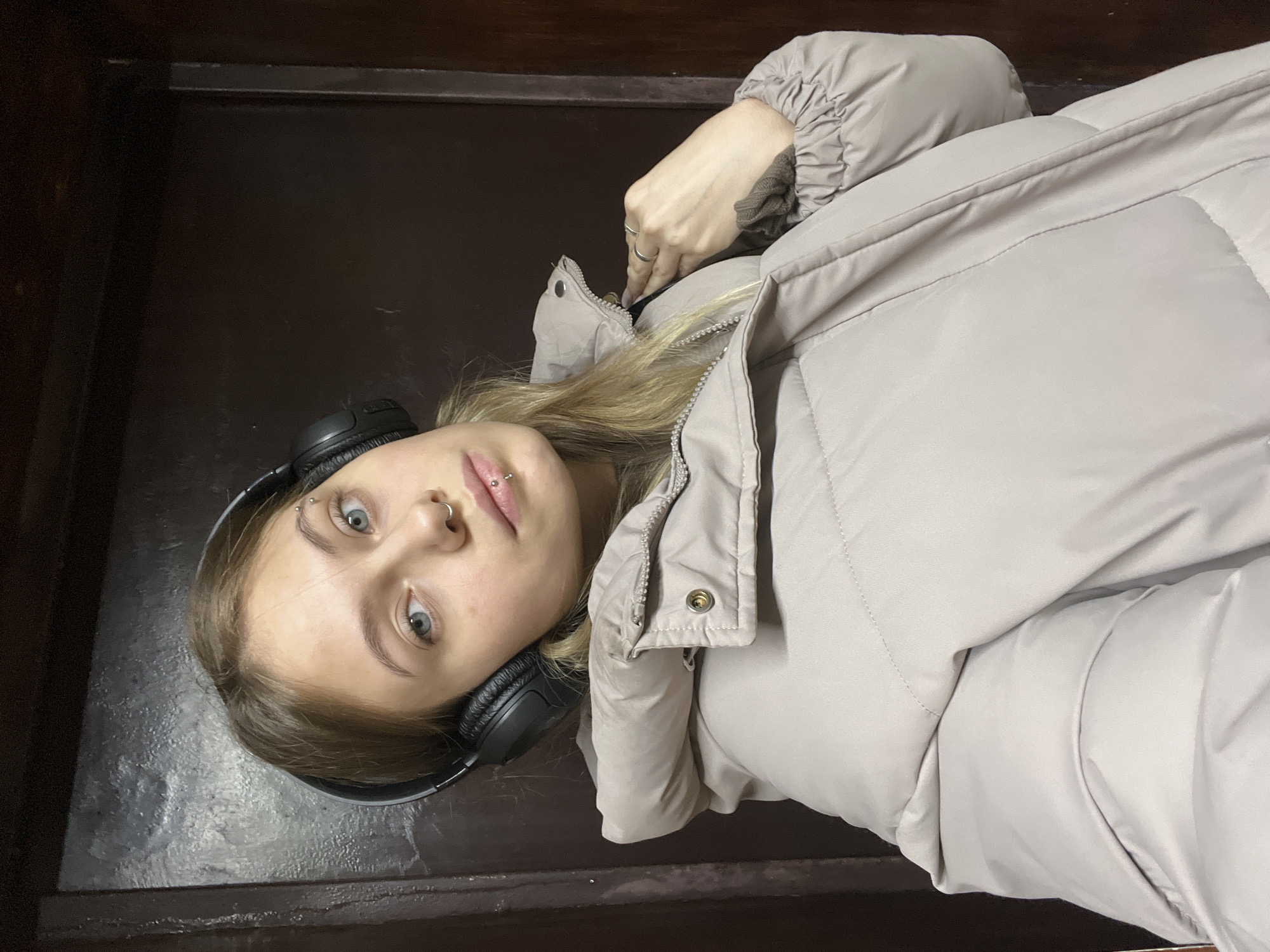 An indoor portrait displays a young female subject positioned against a dark, paneled background. The individual, with light-colored hair and blue eyes, wears black over-ear headphones and a light beige puffer jacket. Facial piercings include a silver ring in the left nostril, a silver labret stud centered below the lower lip, and a silver barbell in the left eyebrow. Two silver rings are present on the ring finger of her right hand, which rests near the jacket's collar. The subject maintains direct eye contact with the camera and exhibits a neutral facial expression. The specific location within the city of Фоки, Russia, is not discernible from this interior setting.