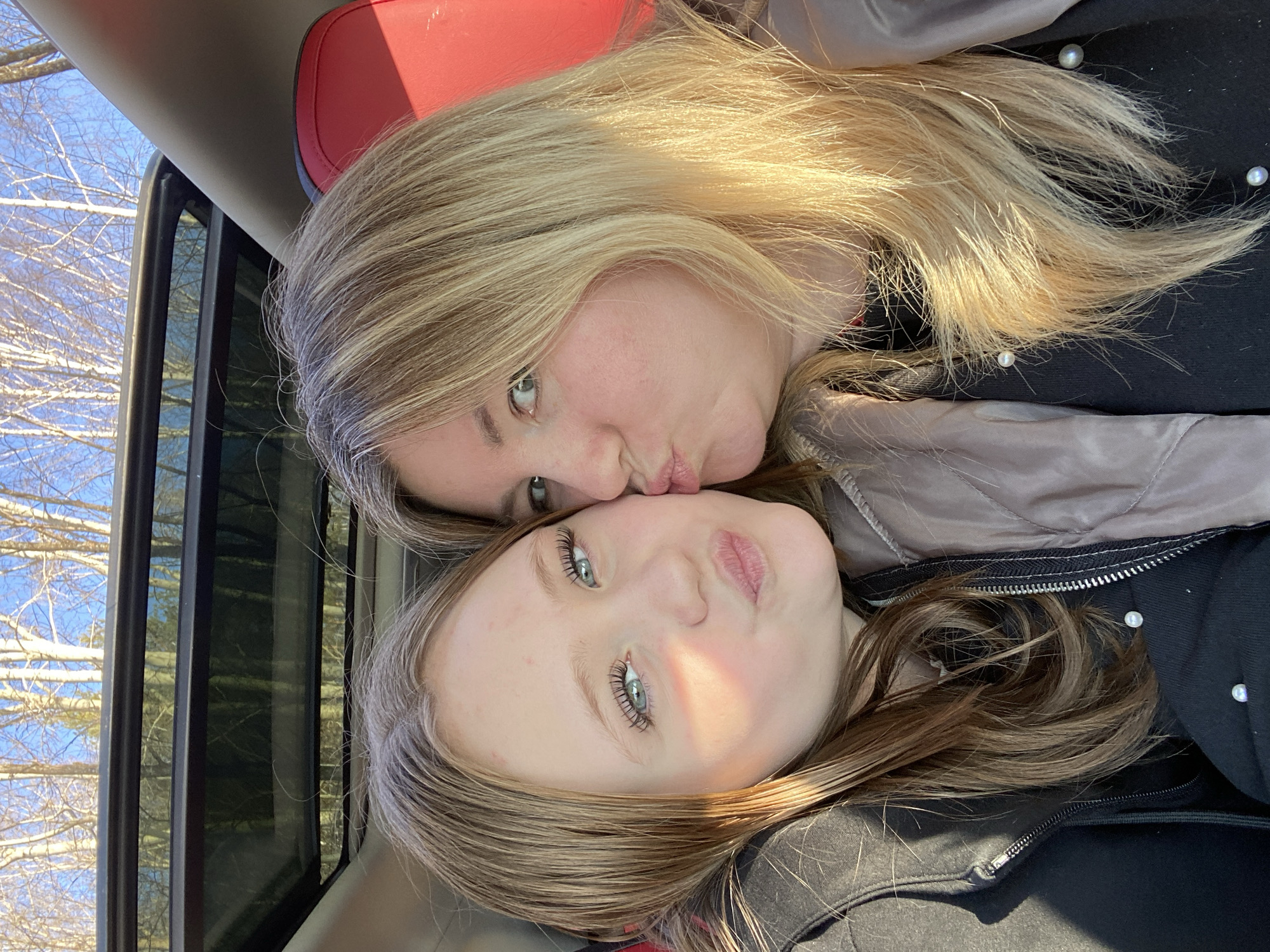 The image captures a close-up, affectionate moment between two females, likely a mother and daughter, taken inside a vehicle. The adult, with long blonde hair, is seen at the top, puckering her lips as if giving a kiss to the younger individual below her. Her green eyes are open and looking towards the camera. The younger girl, with light brown hair and prominent blue eyes, also has her lips slightly puckered, looking towards the camera.
Bright sunlight illuminates their hair and faces, suggesting a clear day. Through the vehicle's window in the background, bare tree branches are visible against a clear blue sky, indicating a season like late autumn, winter, or early spring. The interior of the vehicle shows red upholstery, likely a car seat, in the upper part of the frame, and a dark window frame.
Both individuals are wearing dark, possibly padded, jackets. Several small white spherical beads, resembling pearls, are visible on the dark fabric of their clothing, particularly near the younger girl's shoulder and the older woman's jacket. The scene conveys warmth and affection. The location context is Chaikovsky, Russia. There is no visible text in the media file.