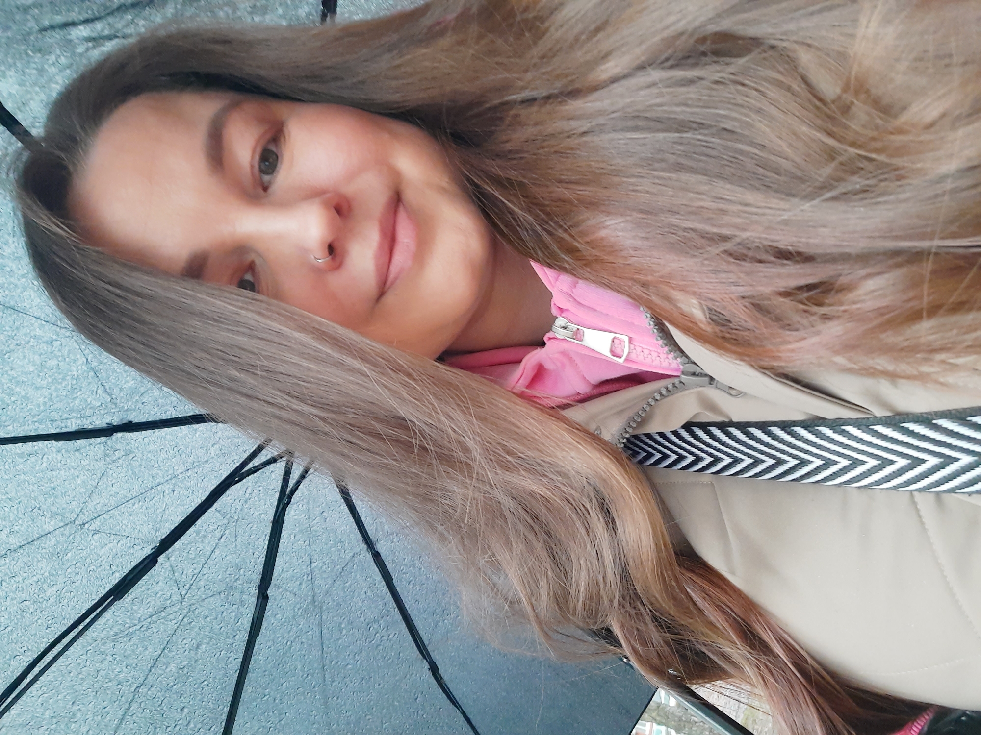 A young woman with long, light brown hair is shown from a close, upward angle. She is smiling slightly and has a nose ring in her left nostril. She is wearing a pink jacket under a beige jacket with a black and white patterned strap. The top of a wet, transparent umbrella with black ribs is visible, indicating it may be raining or recently rained. There is no indication that the image was taken in Izhevsk, Russia.