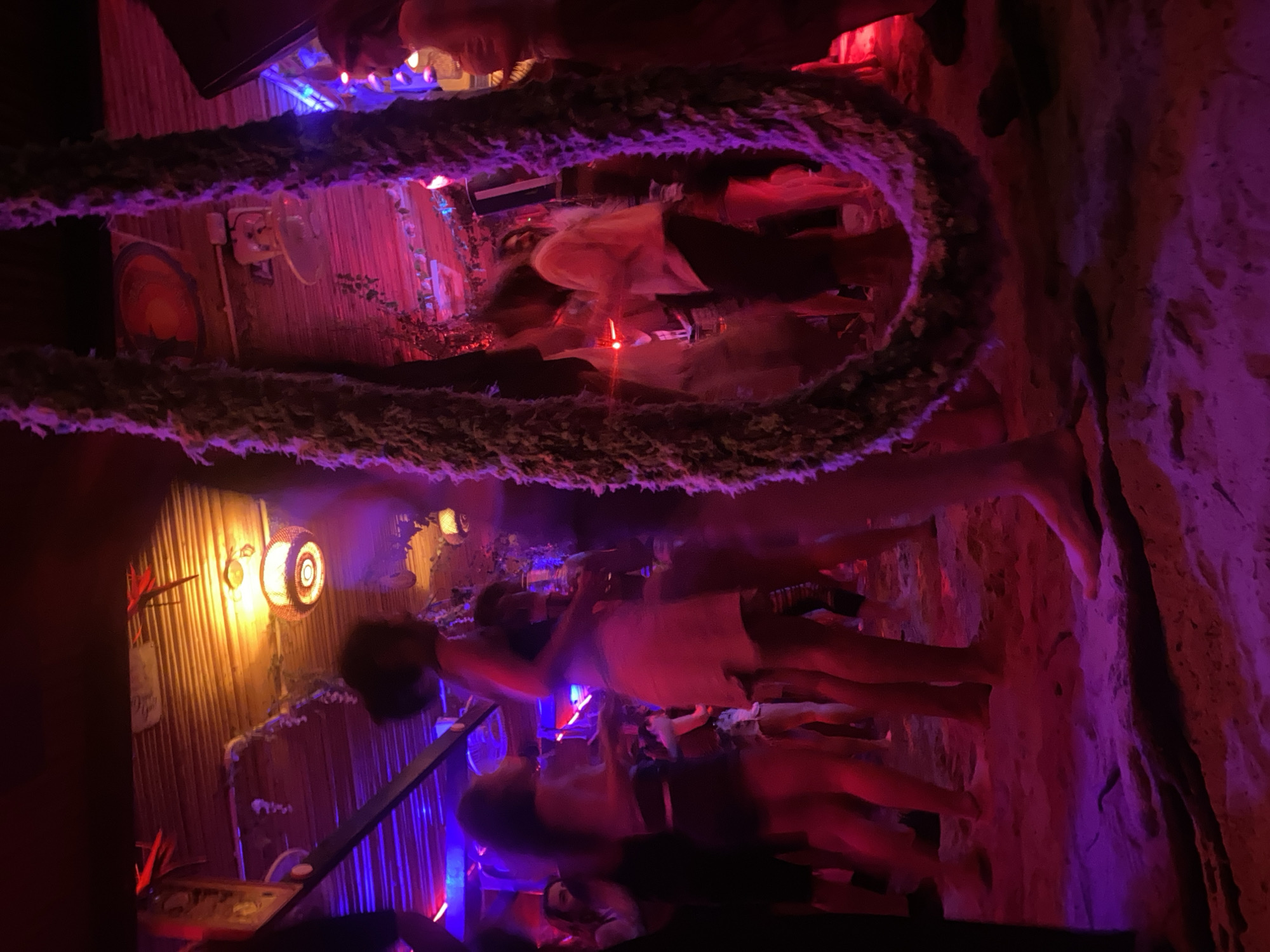 The image depicts a dimly lit interior in Baan Sri Thanu, Thailand, possibly a bar or club, featuring a decorative rope hanging in the foreground. Several people are present, though their actions and interactions are difficult to discern due to the blur and lighting. The background consists of bamboo-style paneling, various light fixtures, and red and blue lights illuminating the scene. A ladder or railing is also visible. A rock-like structure with pink lighting is on the right side of the image.
