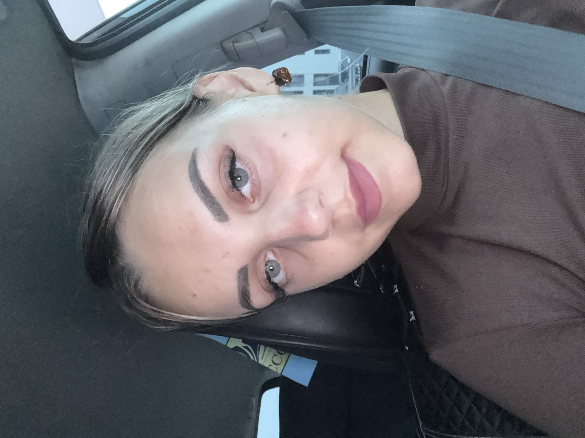 The image is a close-up selfie taken inside a car, likely in the city of Yuzhno-Sakhalinsk, Russia. A young woman with blonde hair pulled back in a bun is the primary subject. She has light blue eyes, defined eyebrows, and is wearing makeup, including mascara and lipstick. Her attire consists of a brown turtleneck sweater. A seatbelt is visible across her chest. In the background, a car seat with a patterned cover and a blue and yellow banner are partially visible. The setting suggests an urban environment, with a blurred view of buildings and a street visible through the car window.