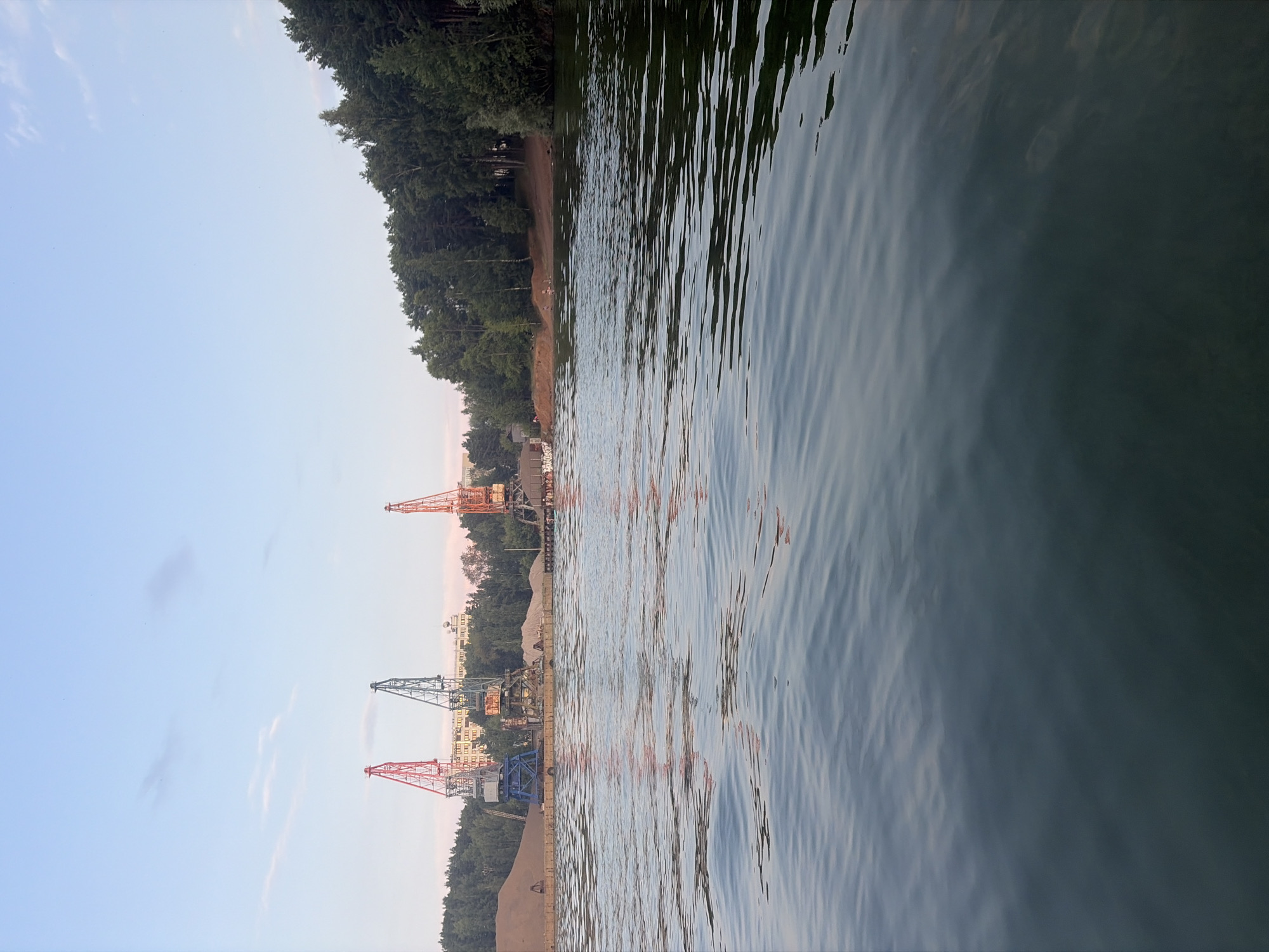 This is a landscape photograph taken from a body of water, likely on the Kama River in Chaikovsky, Russia. In the background, a shoreline is visible with a dense line of green trees. Along the water's edge, there is a dock or pier with several large industrial cranes, some red, blue, and orange, positioned over piles of sand or gravel. Buildings and other industrial structures can be seen behind the cranes, and a light blue sky with wispy clouds fills the upper portion of the frame. The water in the foreground is calm with gentle ripples, reflecting the colors of the sky and the cranes.