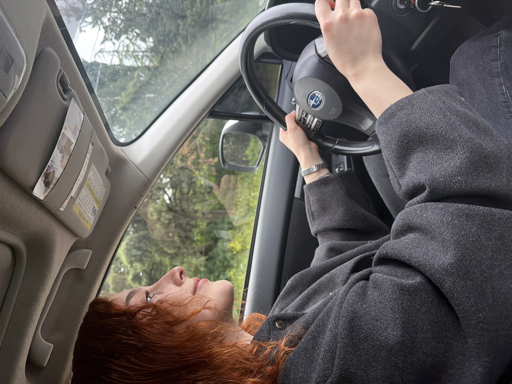 A fair-skinned person with red hair is reclining in the driver's seat of a Subaru vehicle. The person is wearing a dark gray wool coat and a silver-colored wristwatch. The car's steering wheel is held in their hands. Through the car windows, foliage can be seen. The location is not possible to determine as the image does not provide enough information to identify the city as Tbilisi, Georgia.