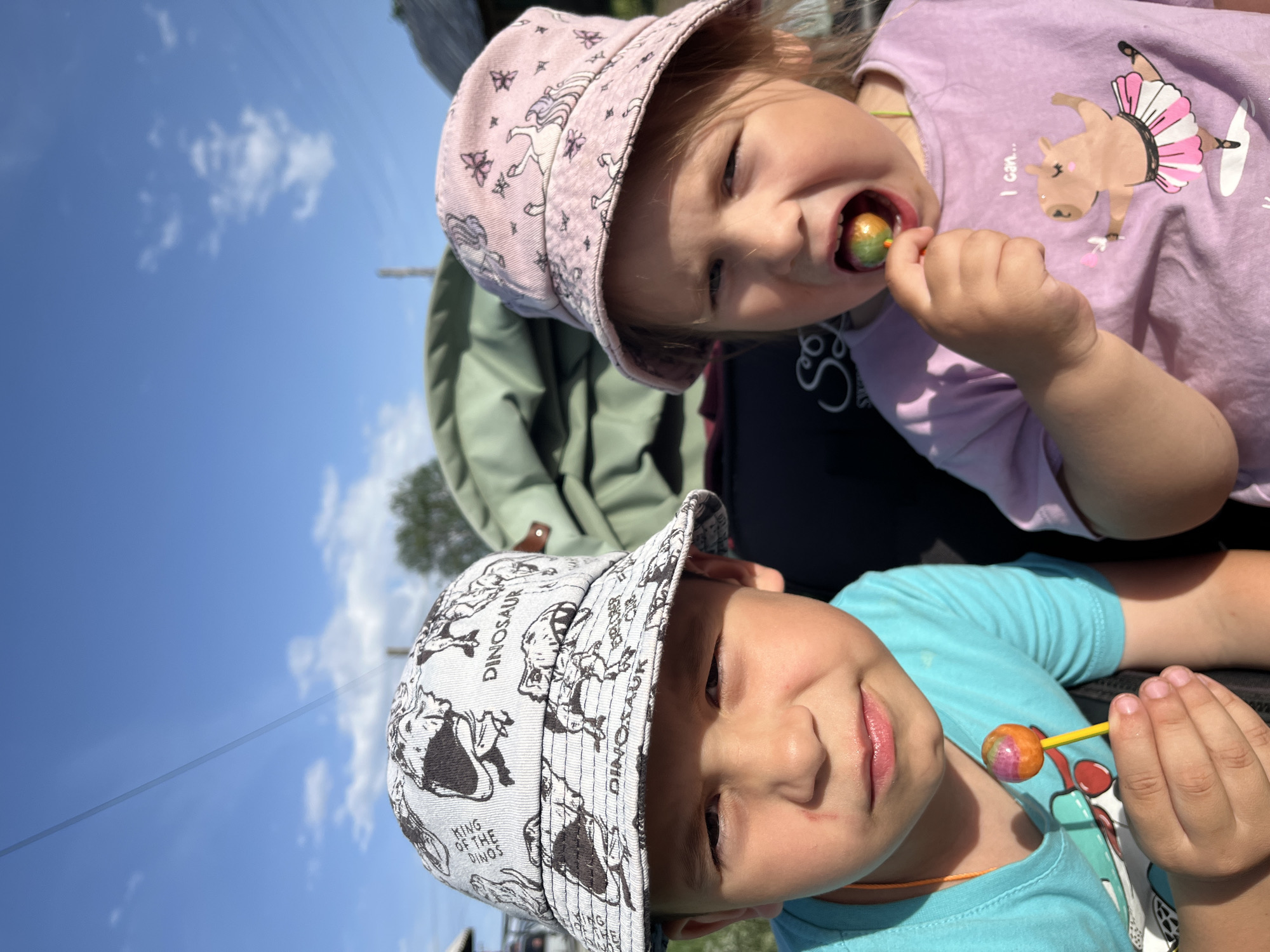 Two young children are the focus of the image, likely taken outdoors in Chaikovsky, Russia, given the prompt. The child in the foreground is a boy wearing a light blue T-shirt and a bucket hat patterned with dinosaurs, some labeled "DINOSAUR" and "KING OF THE DINOS." He is smiling slightly and holding a lollipop. Behind him and slightly above is a girl wearing a pink T-shirt with a dancing pig design and a pink bucket hat adorned with unicorns. She is actively eating a lollipop. Both children appear to be sitting, possibly in a stroller, as there is a green backpack or stroller seat visible in the background. The sky is blue with scattered white clouds. A power line cuts diagonally across the sky in the upper left.