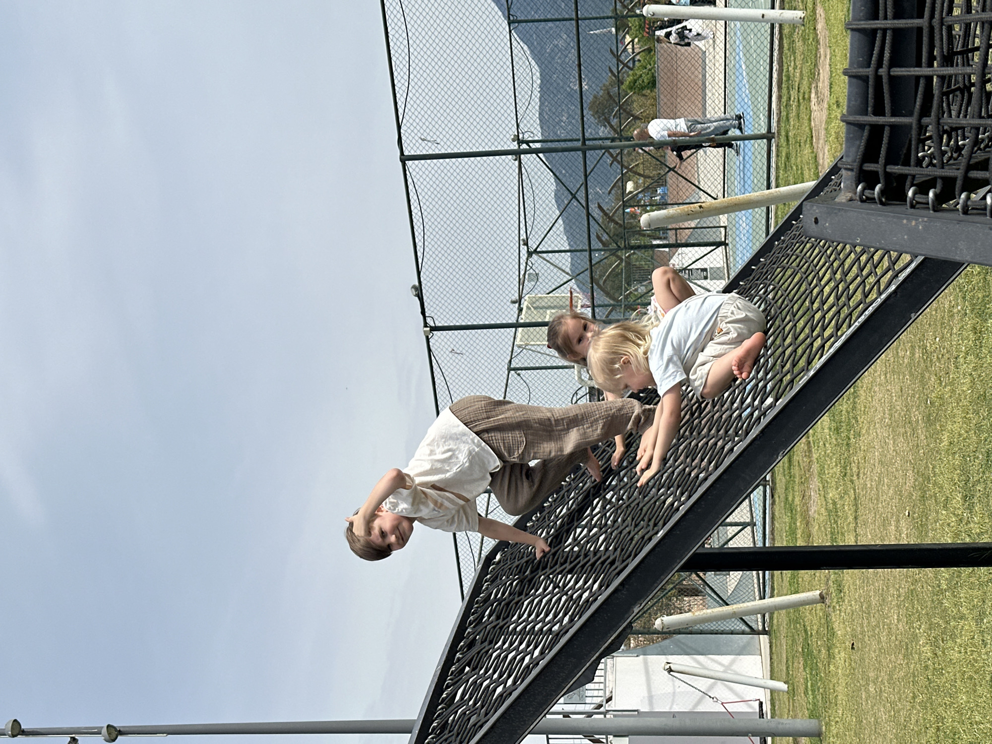 The photograph captures three children playing on a net climbing structure in Fethiye, Turkey. One child, a boy with brown hair, is climbing down the net towards the foreground. He is wearing a white shirt and brown pants. Two other children, a girl and a younger boy, are climbing up the net behind him. The girl has blonde hair, wearing a white shirt and shorts, and is crawling up to the right. The net structure is black and angular, and it is set against a backdrop of green grass, a chain-link fence, and buildings in the distance. The sky is visible above the structure, appearing light blue and somewhat overcast. In the background, through the fence, there are indications of a sports area, possibly a tennis court, with other people present. The photograph is taken during the day, likely in an outdoor play area or park within the city of Fethiye.