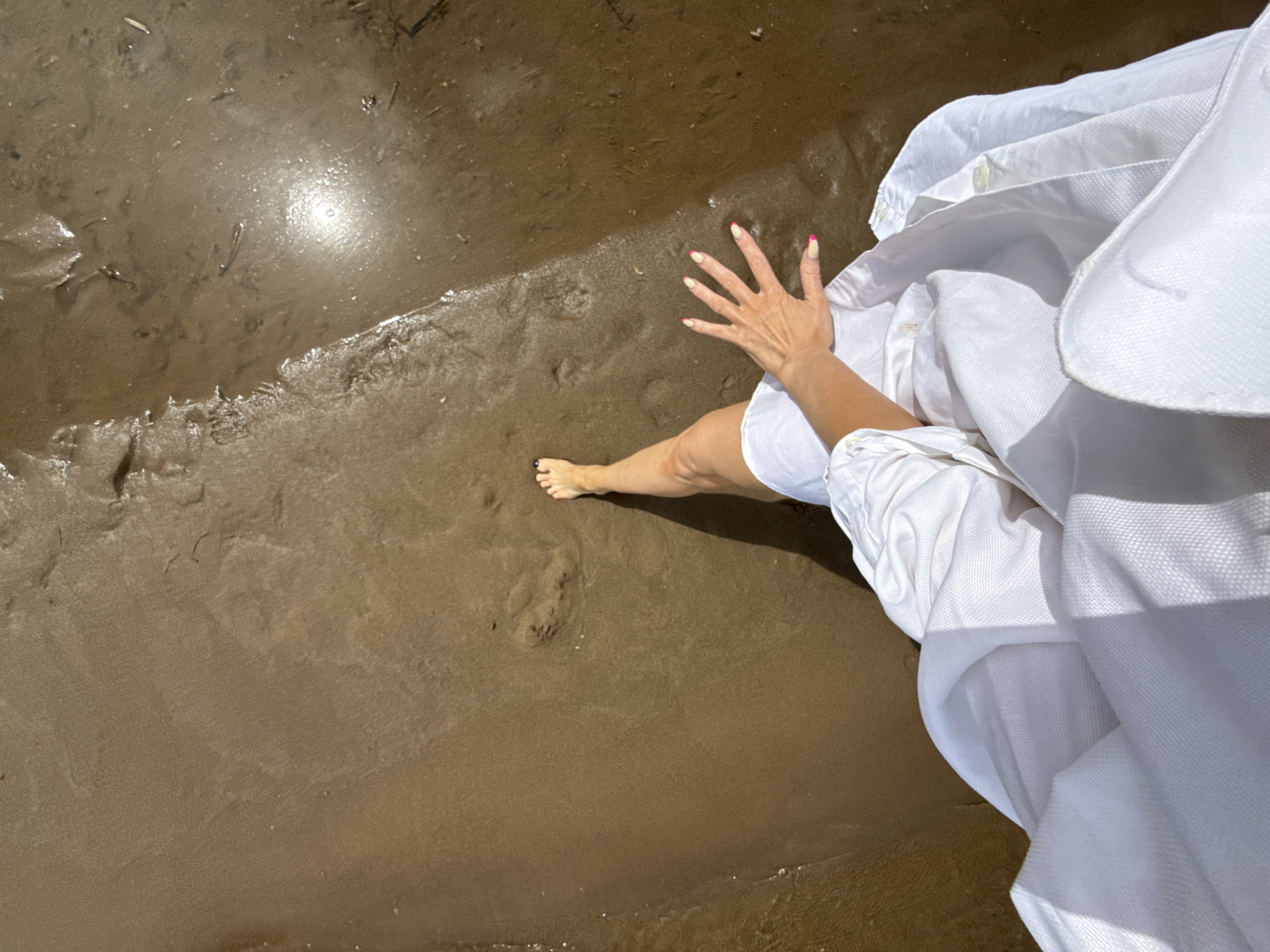 A woman in a white garment stands on a wet, brown sand beach. Her left foot is visible near a small footprint in the sand. Her right hand is outstretched, resting on the wet sand. The scene is illuminated by sunlight, reflecting off the wet sand. It may be located in the city of Chaikovsky, Russia.