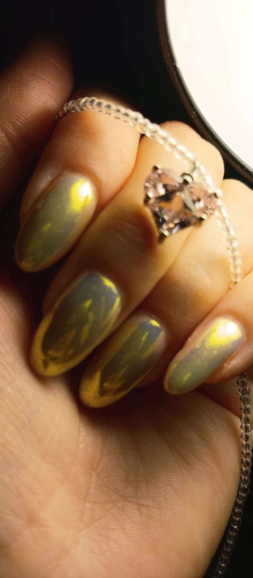 A hand holds a necklace with a heart-shaped pendant. The necklace chain is made of small, clear beads. The pendant appears to be a pinkish-purple stone or crystal set in a silver or light-colored metal setting. The hand has long, almond-shaped fingernails with a metallic, iridescent nail polish that shifts between green, yellow, and gray tones. The background includes a dark area and a brighter, light-colored area, possibly suggesting an indoor setting with mixed lighting. The location is unknown.