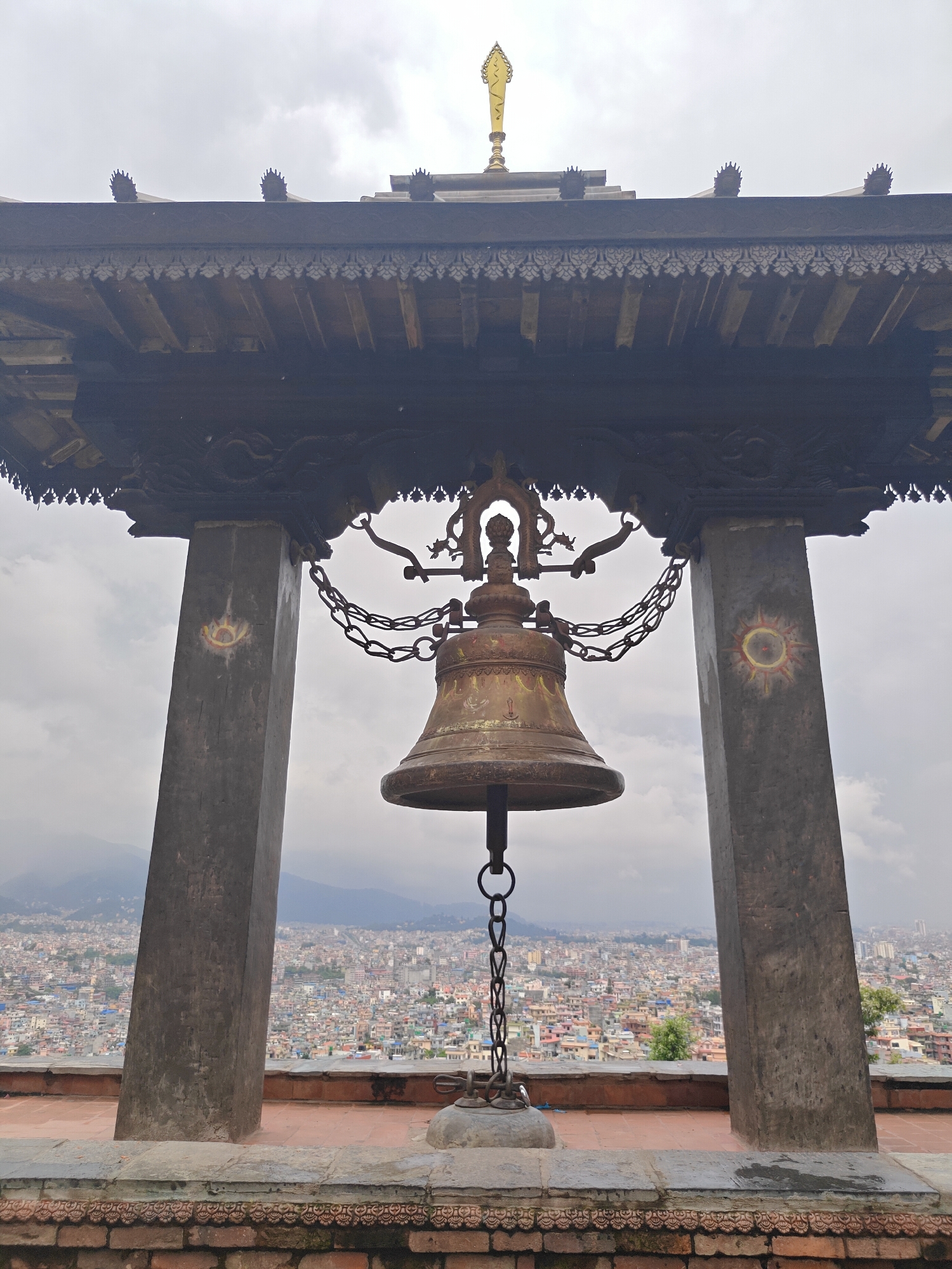 A large, bronze bell hangs from a wooden structure supported by two stone pillars, overlooking the city of Kirtipur, Nepal. The bell is intricately designed with chains connecting it to the overhead structure. The pillars are adorned with circular, sun-like symbols. The city sprawls across rolling hills under a cloudy sky. The foreground shows a tiled platform and a brick wall with decorative carvings. A golden spire is visible atop the wooden structure.
