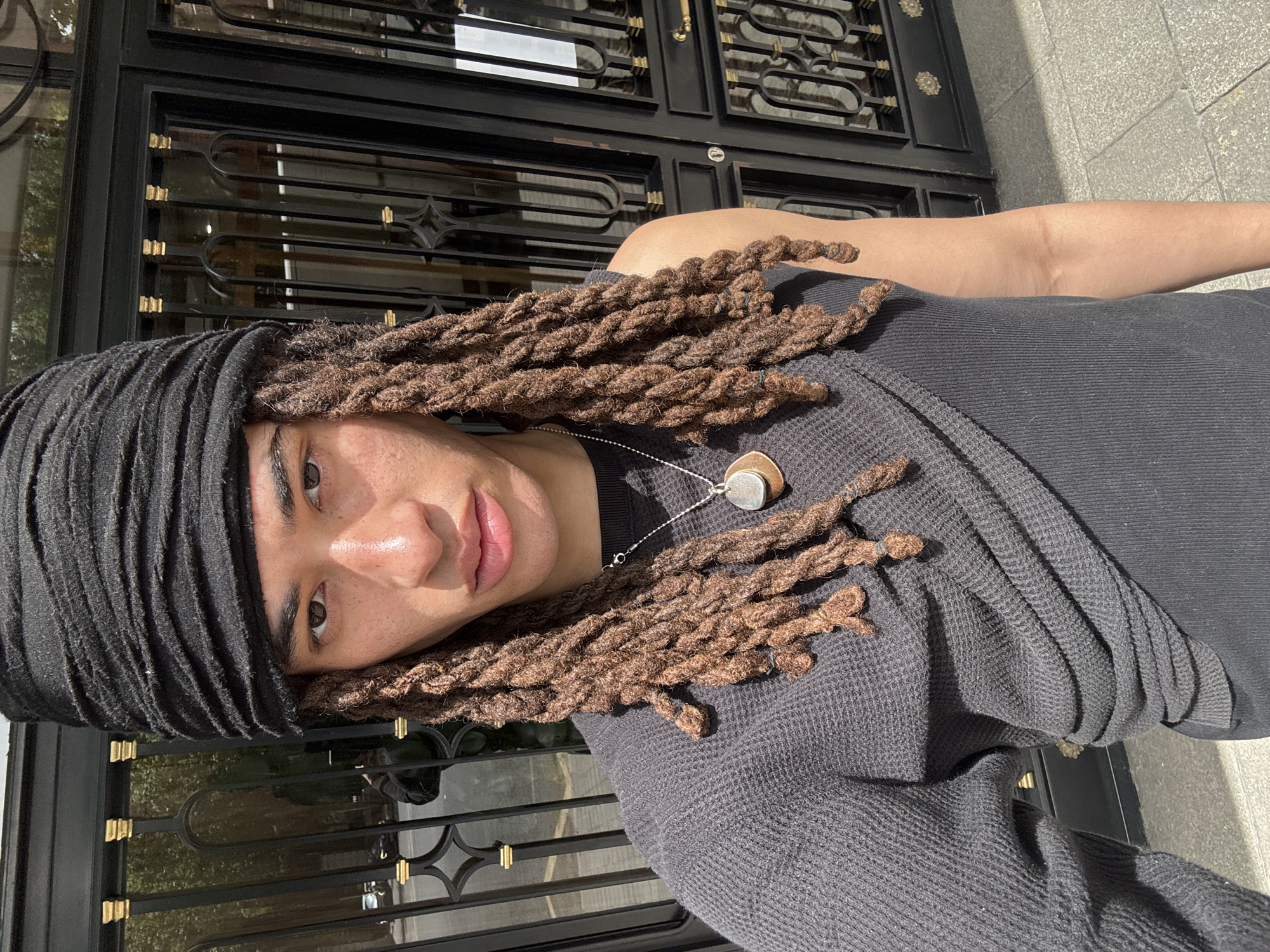 The image captures a person in a close-up, selfie-style portrait shot outdoors in Madrid, Spain. The individual has brown dreadlocks, many of which are visible flowing over their left shoulder and chest, and wears a black, textured headwrap that covers part of their forehead. They are dressed in a black waffle-knit long-sleeved top and accessorized with a silver chain necklace featuring two pendants: one metallic silver and the other a natural, brown material, possibly wood or stone. The person looks directly at the camera with a neutral to slightly serene expression, and visible freckles dot their nose and cheeks.
The background is bathed in bright sunlight, indicated by strong shadows and highlights. Behind the individual is an ornate black metal gate or fencing with decorative golden accents, suggesting an upscale or historically styled building. Reflections can be seen in the background panels or windows behind the gate. To the right, glimpses of light-colored paving stones suggest a street or pathway. No discernible text is visible in the image.