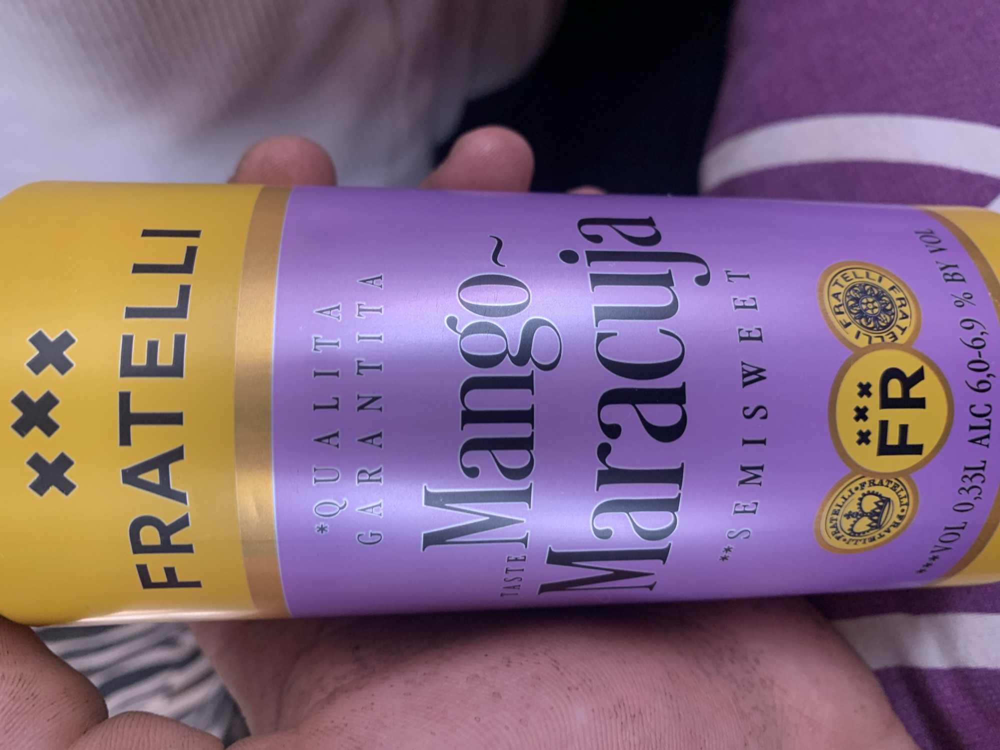 The image shows a close-up of a can of "FRATELLI Mango Maracuja" drink. The can has a yellow top with "FRATELLI" written in large black letters and three black crosses above it. The main body of the can is purple with the words "*QUALITA GARANTITA" in white text. Below this, the flavor "Mango~ Maracuja" is prominently displayed in a larger, serif font. At the bottom of the purple section, "**SEMISWEET" is written. There are also two circular logos, one with a crown and the other with "FR" inside a yellow circle. The bottom of the can shows text indicating "VOL 0,33L ALC 6,0-6,9 % BY VOL". A person's hand, which appears slightly dirty, is holding the can. The background is blurred and appears to be a striped fabric. There is no information in the image that could indicate the location is Zhmerynka, Ukraine.