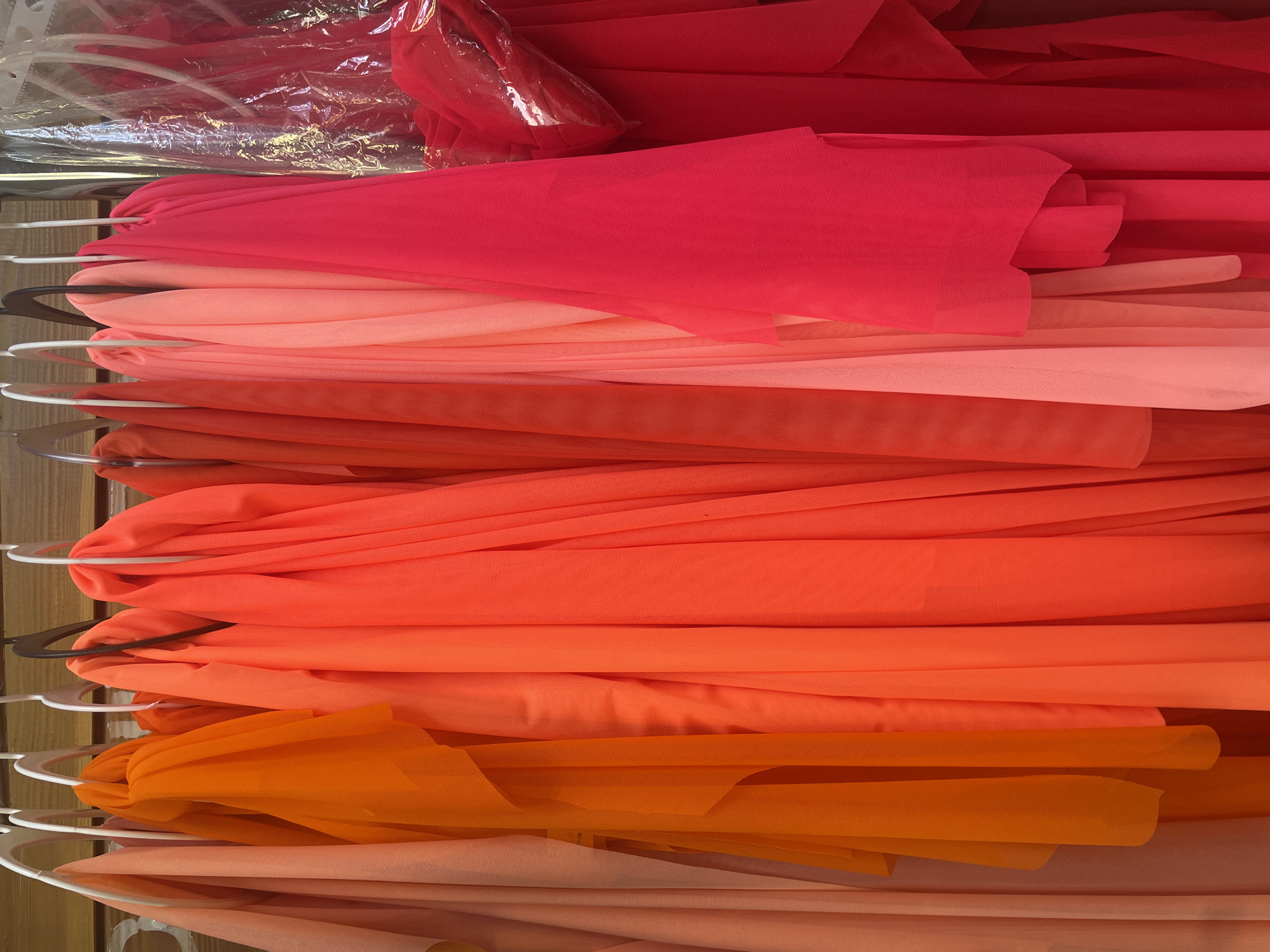 A rack of brightly colored fabrics, primarily in shades of orange, pink, and red, is displayed. The fabrics are draped and folded on hangers, creating a vibrant and textured visual. The scene appears to be in a retail setting, likely a fabric store or a boutique. There are no people or specific landmarks visible to indicate a city or location.