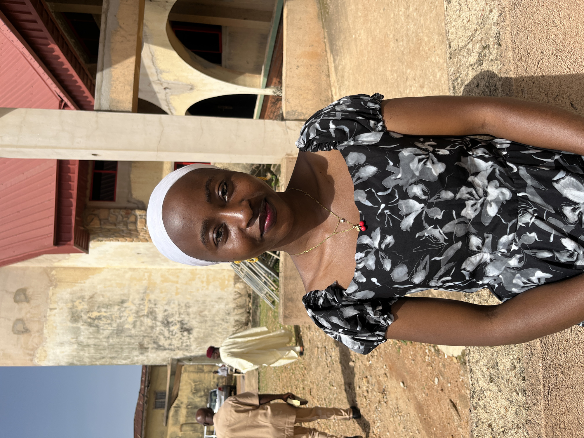 A medium shot shows a young Black woman, head and shoulders, wearing a white hijab, a gold necklace with a cherry pendant, and a black dress with a white floral pattern. She is looking directly at the camera and smiling faintly. She is outdoors, seated in front of a textured, light brown wall, possibly a bench or low structure.
In the background, a pale yellow building with a red roof is visible. The building has arched windows and columns. To the left, two men are walking away from the camera on a gravelly path. One man wears a tan outfit and carries a book. The other wears a traditional light yellow robe and a red cap. A white car is parked behind them. The sky is clear and blue.
The overall impression is of a sunny day in an outdoor location, possibly a place of worship or a historical site in Bukuru, Nigeria. The lighting suggests it is daytime, likely mid-morning or afternoon given the shadows.