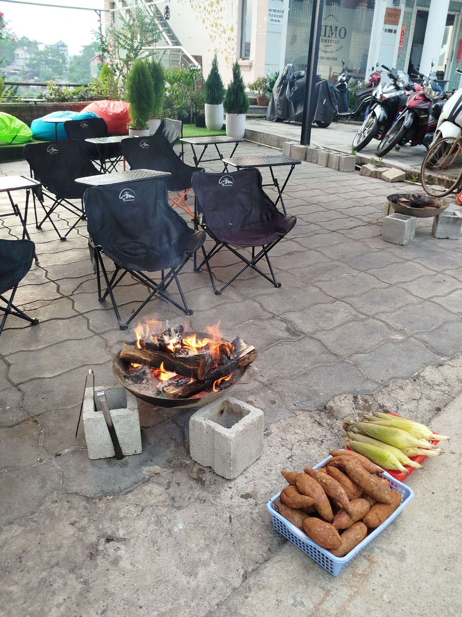 The image shows an outdoor scene in Da Lat, Vietnam, featuring a gathering for a fire pit cooking session. Several black folding chairs with small tables are arranged around a burning fire pit, elevated on cinder blocks. A basket of sweet potatoes and a tray of corn on the cob are set nearby. In the background, there are motorbikes parked near a building with a sign that reads "IMO HOMESTAY". The location appears to be a patio or rooftop area with a view of the surrounding city. The OCR also shows the text: "CHO THUE XE Ô TÔ TỪ LÀI VÀ XE MÁY" "khoản khắcủa ban" which could be a sign offering car and motorcycle rentals.