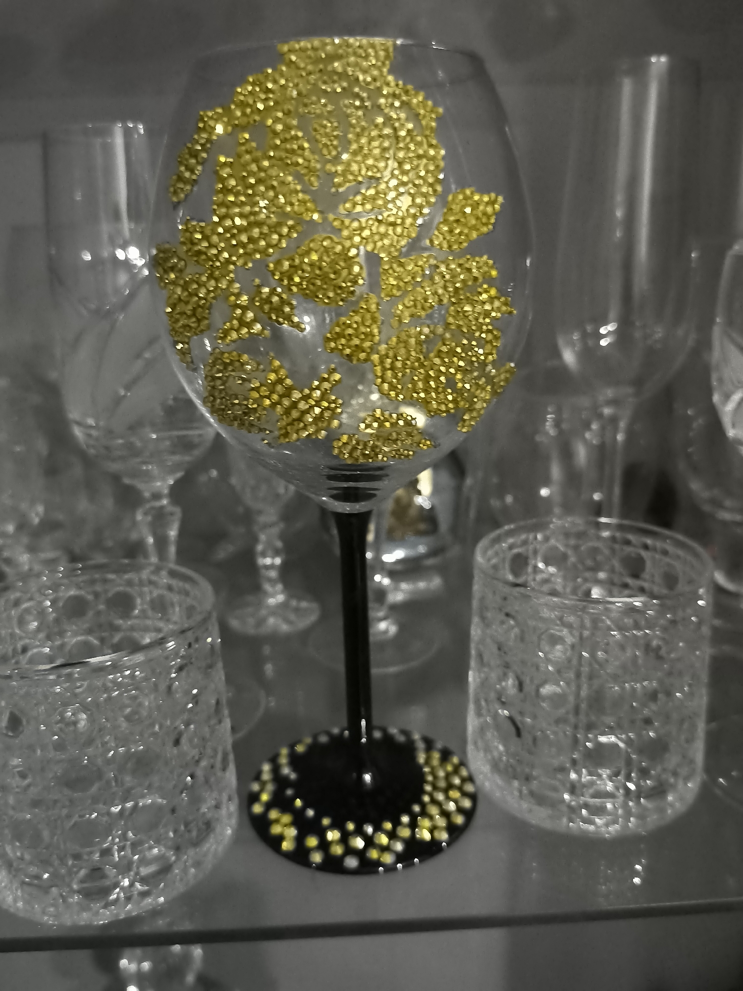 The image shows a close-up of glassware with a focus on a wine glass decorated with gold rhinestones. The wine glass has a black stem and base, also adorned with gold and white rhinestones at the bottom. To the left and right of the wine glass are two short, clear tumblers with an intricate patterned texture. In the background, other clear glasses are visible, suggesting they are part of a collection or display. The lighting is somewhat dim, creating reflections on the glass surfaces. The scene is static, with no people or actions depicted. The setting appears to be indoors, possibly a shelf or display case in a home or a store. There is no indication of the city of Klaipėda, Lithuania, in the image or its context.