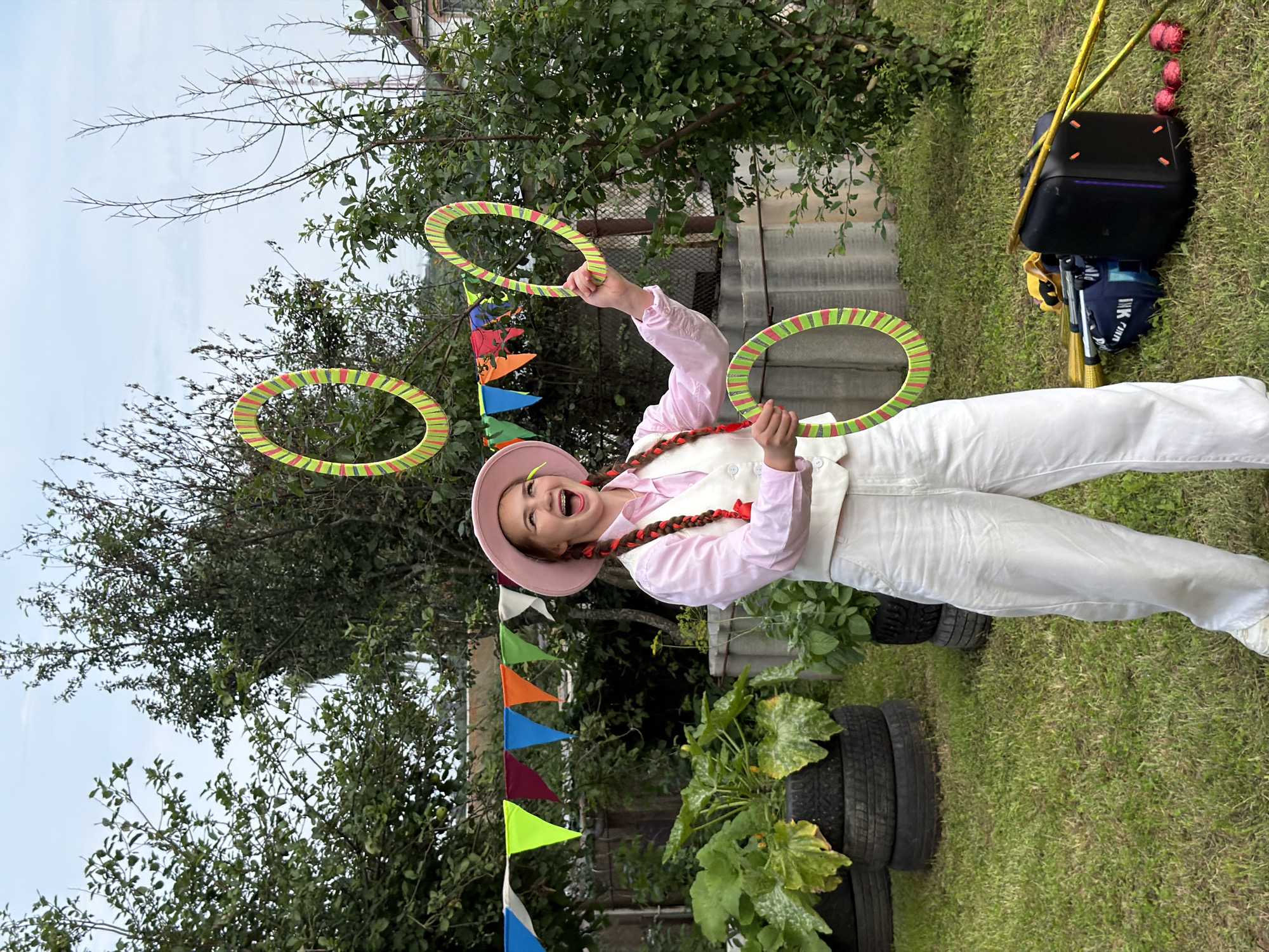 A young person is juggling three colorful rings in an outdoor backyard setting. The person is wearing a pink hat, a pink button-up shirt, a white vest, and white pants, with braided hair adorned with red ribbons. The backdrop consists of trees, a fence with colorful bunting, and some gardening plants growing out of tires. Various props, including a black suitcase, bags, and yellow sticks, are scattered on the grass. The scene appears to be in the city of Chaikovsky, Russia.