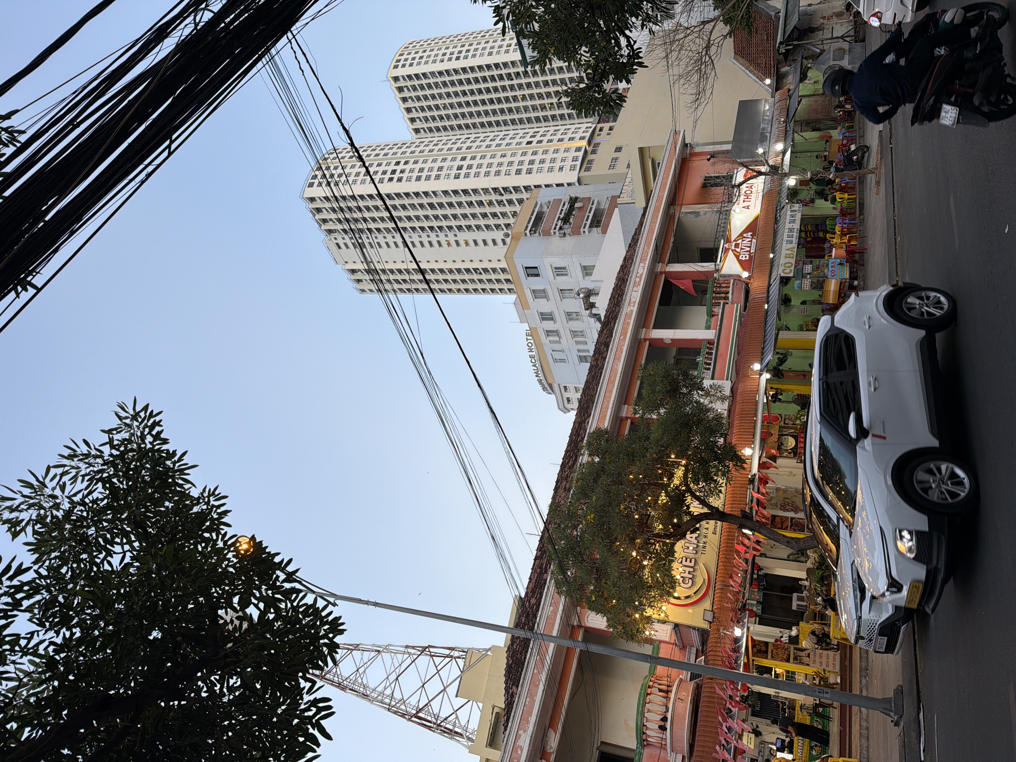 The image depicts a street scene in Nha Trang, Vietnam, with a focus on a row of buildings and high-rise apartments under a clear sky. In the foreground, two white cars are parked on the street, and a person on a motorcycle is riding past. The buildings in the middle ground appear to be businesses with outdoor seating and signage in Vietnamese. One sign reads "CHÉ MÃ" and another says "BIVINA ĂN THOẠI". The architecture is a mix of traditional and modern styles, with a pink-colored building featuring balconies in front of taller, modern apartment buildings. Overhead, a dense network of electrical wires crisscrosses the sky, and a tall radio tower is visible in the distance. Lush green trees with decorative string lights add to the urban landscape.
