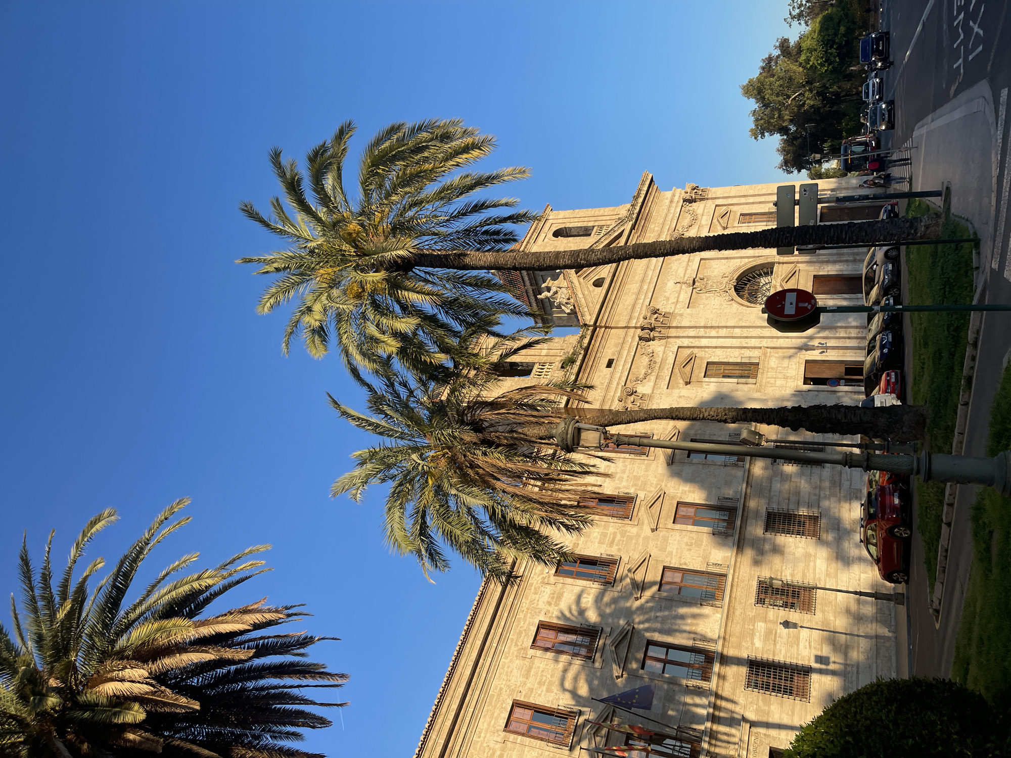 The image shows a sunny day in València, Spain, with a view of the Diputació de València (Valencia Provincial Council) building partially obscured by palm trees. The building is a large, light-colored, multi-story structure with arched windows and ornate architectural details. Several palm trees are in the foreground, their fronds extending towards the clear blue sky. A street runs alongside the building, with parked cars and a traffic sign visible. People are walking on the sidewalk. The overall scene presents a combination of urban environment and natural elements.