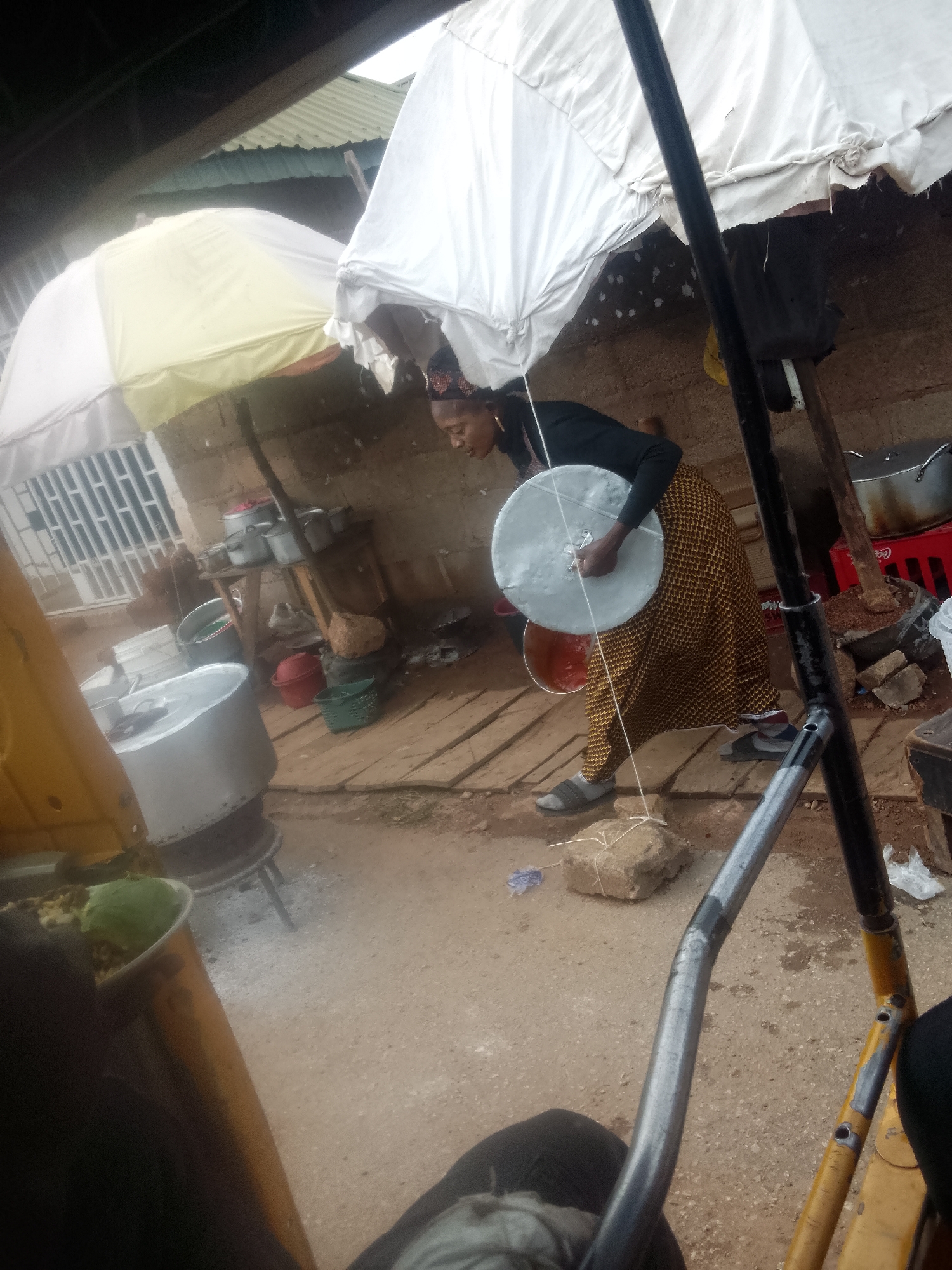 The image, likely taken in Bukuru, Nigeria, shows a woman carrying a large, round, silver metal disc, possibly a lid for a cooking pot. She is wearing a long, patterned brown and gold skirt, a black long-sleeved shirt, and a head wrap. She is standing in a dirt area near a wooden platform. Behind her are various pots, containers, and kitchen equipment, indicating a food preparation area, possibly a street food vendor's setup. Two large white umbrellas offer shade above her and the cooking area. In the foreground are parts of a yellow vehicle frame, possibly a motorcycle or tricycle taxi. A partially visible pot on a stand suggests the presence of an active cooking process nearby.