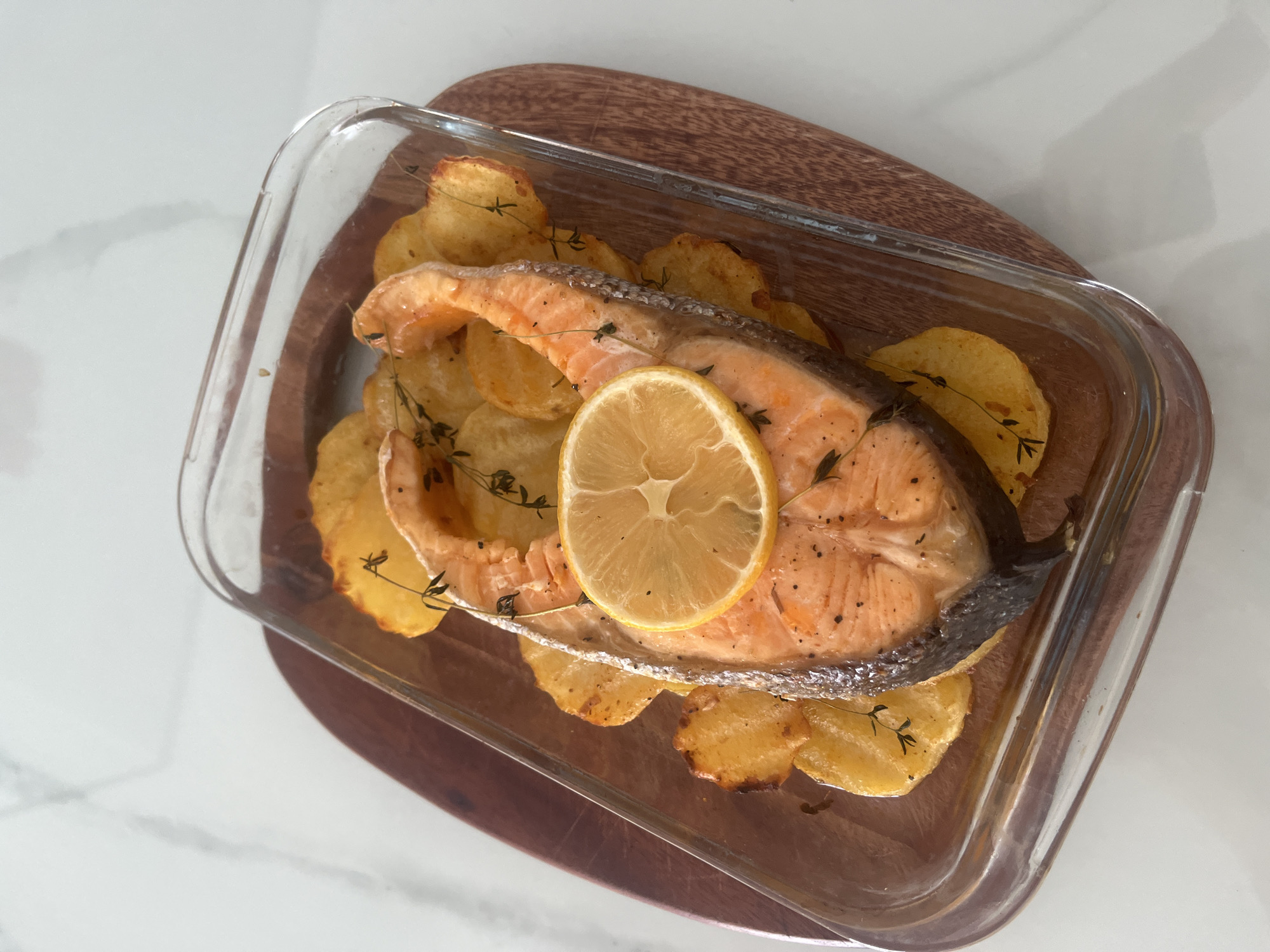 A high-angle shot depicts a cooked salmon steak with a lemon slice on top, garnished with thyme sprigs, resting on a bed of roasted potato slices within a clear glass baking dish. The baking dish sits on a wooden cutting board, which is placed on a white, possibly marble, countertop. The overall composition is bright and well-lit, suggesting a well-lit indoor environment. There is no indication of Bac Nha Trang Ward, Vietnam.
