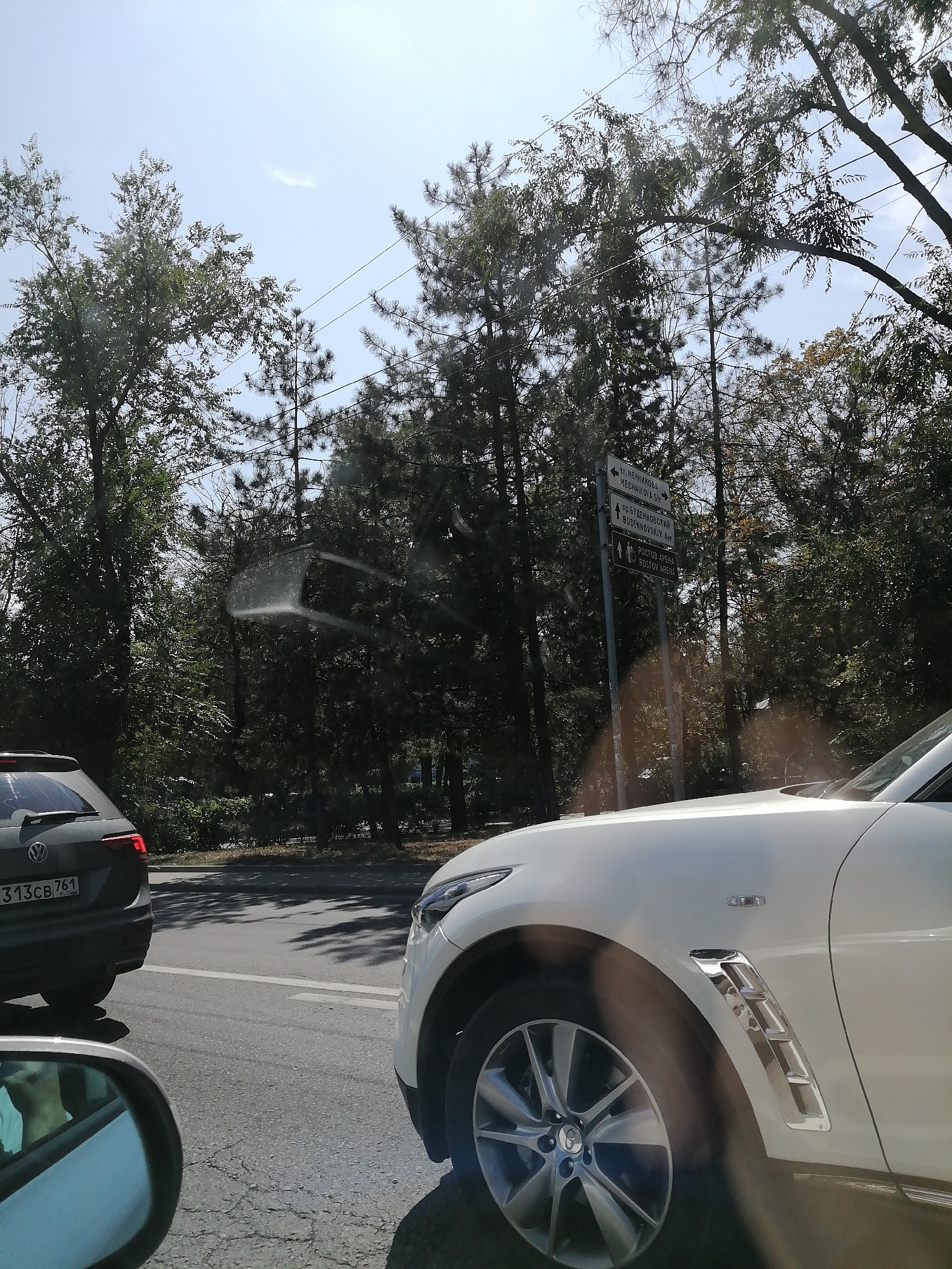 The image captures a sunny day in Rostov-on-Don, Russia, with several cars on a paved road surrounded by dense trees. In the foreground, a white car is prominently displayed, with its front wheel and side panel visible. To the left, a dark Volkswagen SUV is also on the road, its rear bumper and license plate (partially visible as "313CB 761") indicating its presence. A street sign is visible in the middle ground, pointing towards "ул. Мечникова" (Mechnikova Street) and "пр. Буденновский" (Budennovsky Prospekt), and also indicating "РОСТОВ АРЕНА ROSTOV ARENA". The sky is clear with a few faint clouds, and the sun casts shadows on the road. The overall scene suggests a typical urban environment with traffic and greenery.