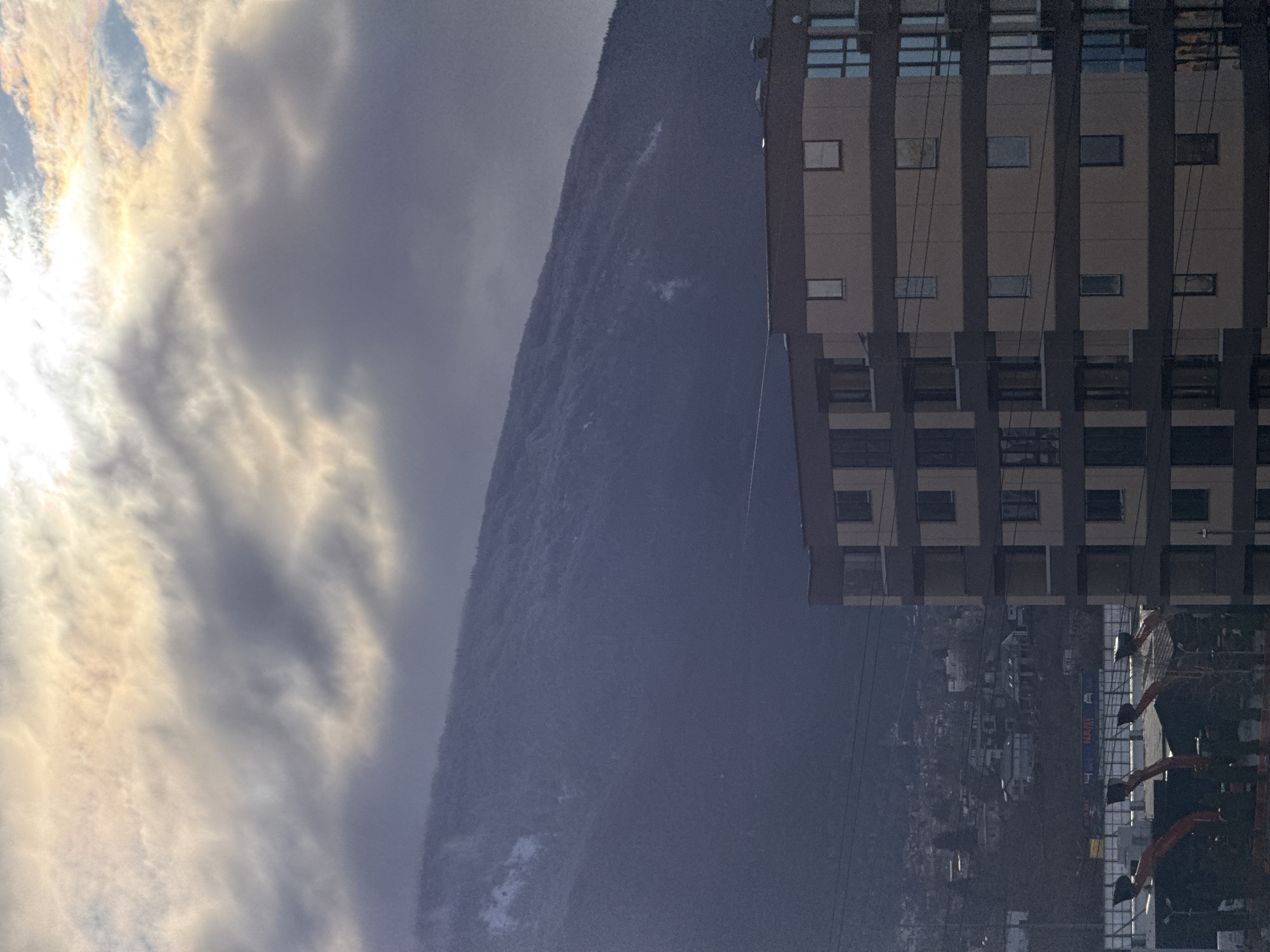 The image depicts a winter or late autumn scene in Sofia, Bulgaria, with the Vitosha mountain range dominating the background. The sky is largely overcast with thick, dark grey clouds, though a bright, diffuse light source, presumably the sun, is visible breaking through the clouds at the top center.
In the foreground, on the right side, a modern multi-story residential building occupies approximately the lower half of the frame. It features a light beige or pale yellow facade with darker brown horizontal bands and vertical frames, suggesting balconies or architectural elements. Multiple rectangular windows are visible, some reflecting the overcast sky.
Below the residential building and to its left, partially obscured, are several large pieces of orange and black machinery, possibly snow-making equipment or construction vehicles. Further into the midground, a densely populated residential area with various buildings and houses is visible along the lower slopes of the mountain. Utility lines traverse the midground, crossing over the residential building and the machinery.
The Vitosha mountain range forms the prominent background, rising steeply and appearing dark blue-grey. Patches of snow are visible on its upper slopes, indicating cold weather conditions. The overall lighting is subdued, characteristic of an overcast day. No individuals are visible in the scene.