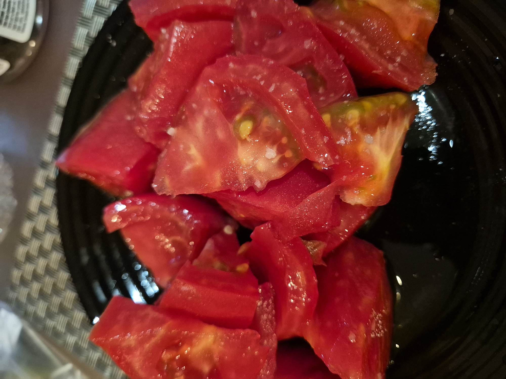 A close-up, overhead shot shows a black plate filled with chopped, ripe tomatoes. The tomatoes are cut into large, irregular pieces, showcasing their juicy interiors and bright red skin. Flecks of salt are visible sprinkled over the tomatoes. The plate rests on a textured placemat. The lighting creates bright highlights on the wet surfaces of the tomatoes, emphasizing their freshness. The background is blurred, with hints of a bottle and other items. The scene appears to be indoors, likely in a kitchen or dining area.