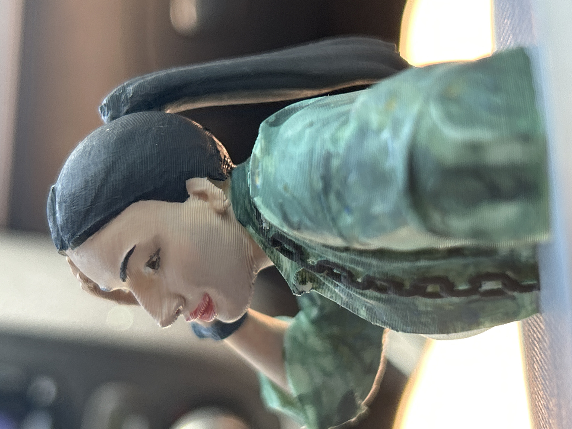The image is a close-up, side profile of a 3D printed figurine of a woman. The figurine has dark hair styled in a ponytail, with bangs that fall over her forehead. She is wearing a green jacket with a camouflage pattern and a dark chain draped around her neck. Her skin tone is light, and she has red lips. One of her arms is raised, with her hand resting on her head. The figurine appears to be placed on a light-colored surface, possibly a table or shelf, with a blurred background that suggests an indoor environment. There is no indication of the city of Pancharevo, Bulgaria in the image.