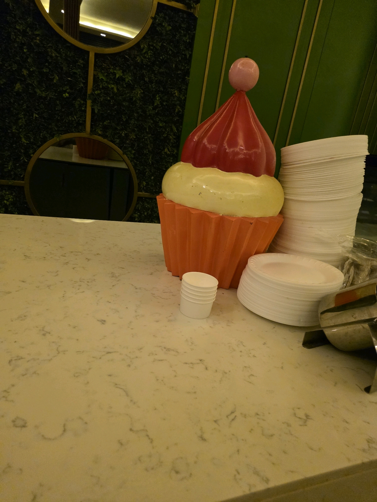 This image depicts a decorative oversized cupcake statue situated on a speckled countertop. The cupcake features a red frosting top with a pink ball ornament, a yellow icing layer, and an orange cupcake liner. To the right of the cupcake, stacks of white paper plates are visible, along with a small stack of white paper cups and a metal serving utensil. In the background, a wall covered in artificial greenery is adorned with two circular mirrors, one above the other. The overall setting appears to be indoors, possibly in a restaurant or event space, and the presence of decorative elements suggests a festive or themed environment. There are no people visible in the image. The city of Bodrum, Turkey, is not depicted or referenced in this image.