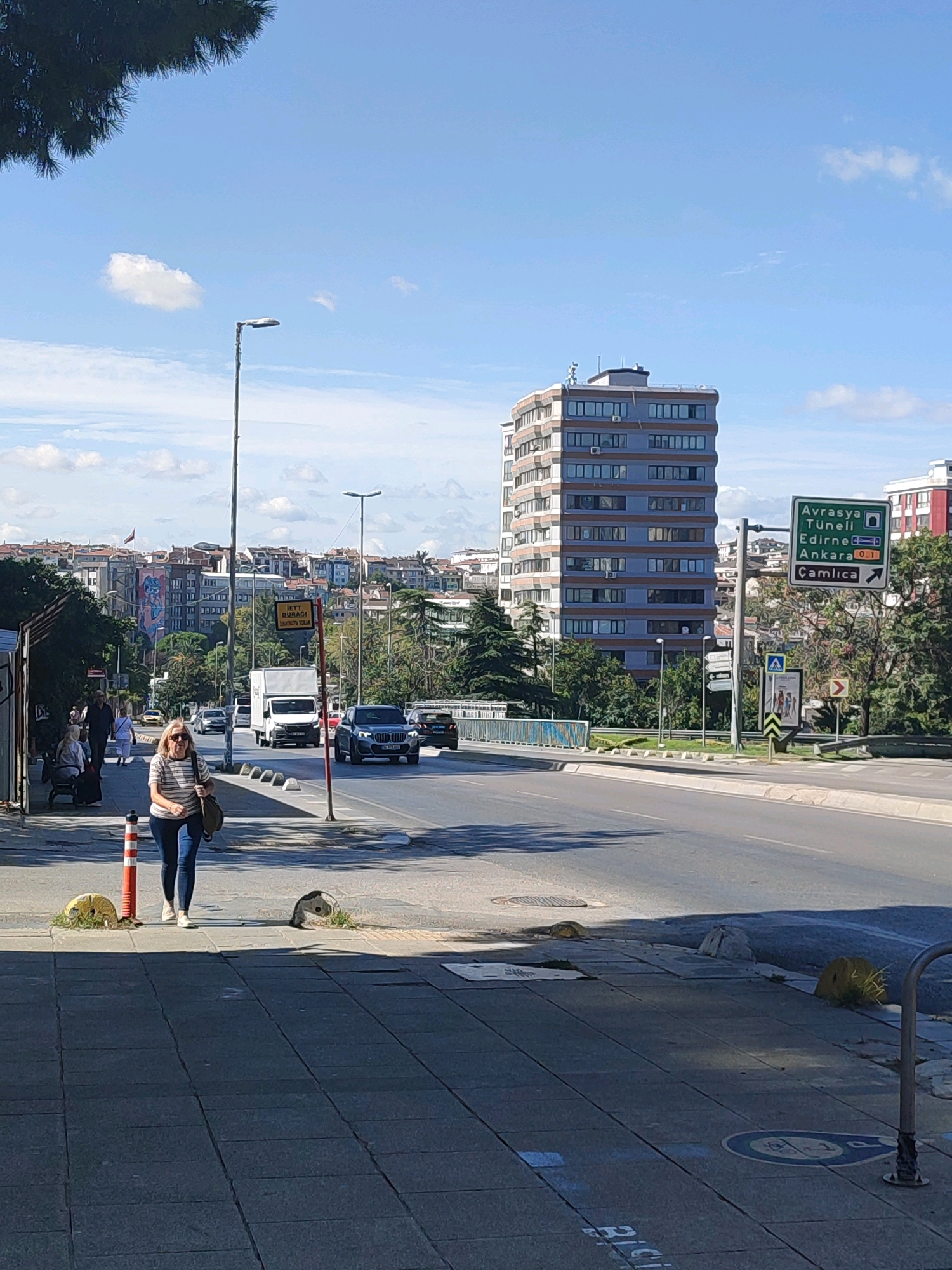 In Kadıköy, Turkey, a woman walks along a sunlit sidewalk next to a busy road. She wears a striped shirt and jeans, carrying a shoulder bag. The sidewalk is made of grey paving stones and is bordered by a road with several cars and a white truck. In the background, a tall, modern building with many windows stands out against a blue sky with scattered clouds. Across the road, a green traffic sign indicates directions to Avrasya Tüneli, Edirne, Ankara, and Çamlıca. The overall scene depicts a typical urban streetscape on a clear day.