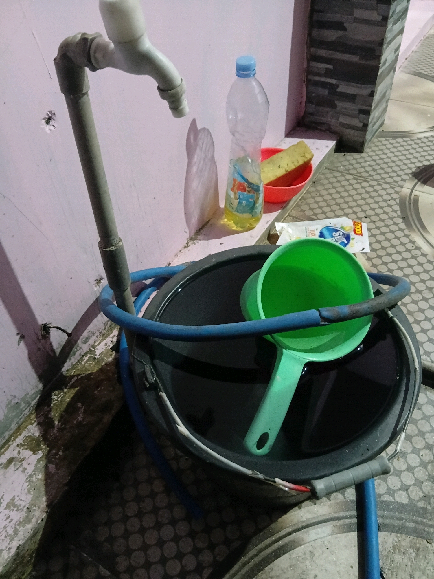 A wide shot shows a faucet connected to a pipe, with a blue hose wrapped around the pipe and leading into a dark bucket filled with liquid. A green ladle rests inside the bucket, partially submerged. To the right of the bucket, a clear plastic bottle with a blue cap is visible, containing yellow liquid and a colorful label. Next to the bottle is a red bowl with a yellow sponge inside. A white packet with blue lettering lies on the ground near the bucket. The scene appears to be outdoors, with a textured tiled floor and a wall made of textured bricks in the background. The lighting suggests it might be evening or a dimly lit area. The overall impression is of a utilitarian setup for water collection or cleaning. The location is described as Cikal, Indonesia.