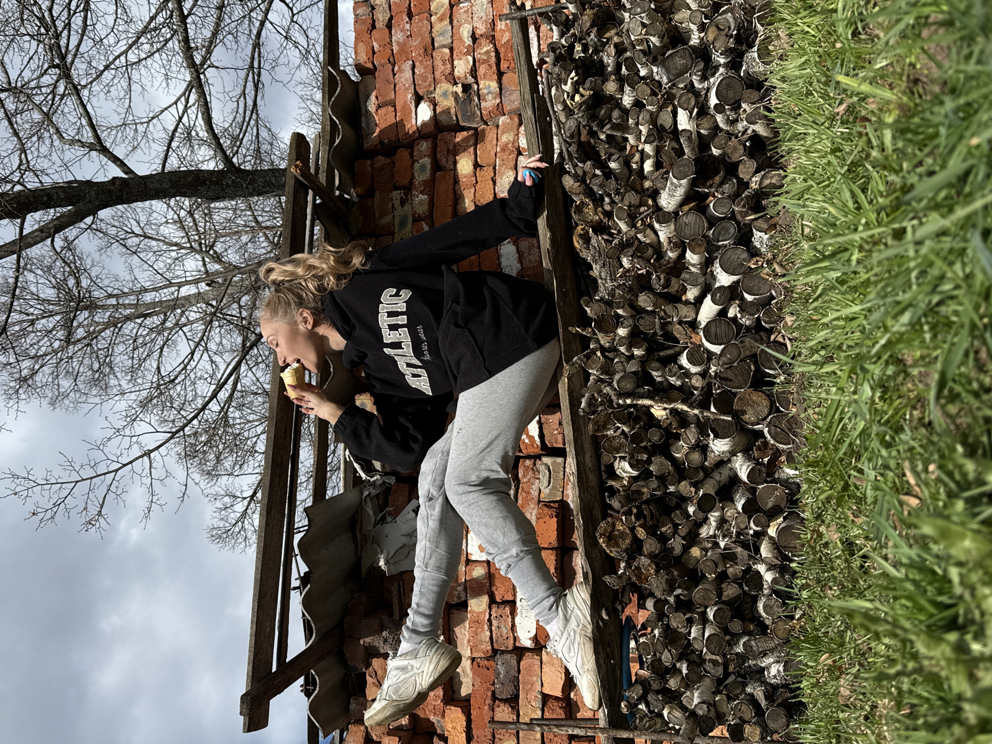 The image depicts a woman eating an ice cream cone in what appears to be a cluttered outdoor setting. She is positioned in front of a stack of firewood and a pile of bricks. A rickety structure made of wooden planks and corrugated metal or roofing material is visible to her left. A bare tree branch extends into the sky above her. The ground to her right is covered in green grass.
Although I am unable to access the location of the city of Rosstani, Russia, the scene appears to be a backyard, a construction site, or a storage area, where materials like firewood and bricks are stored.