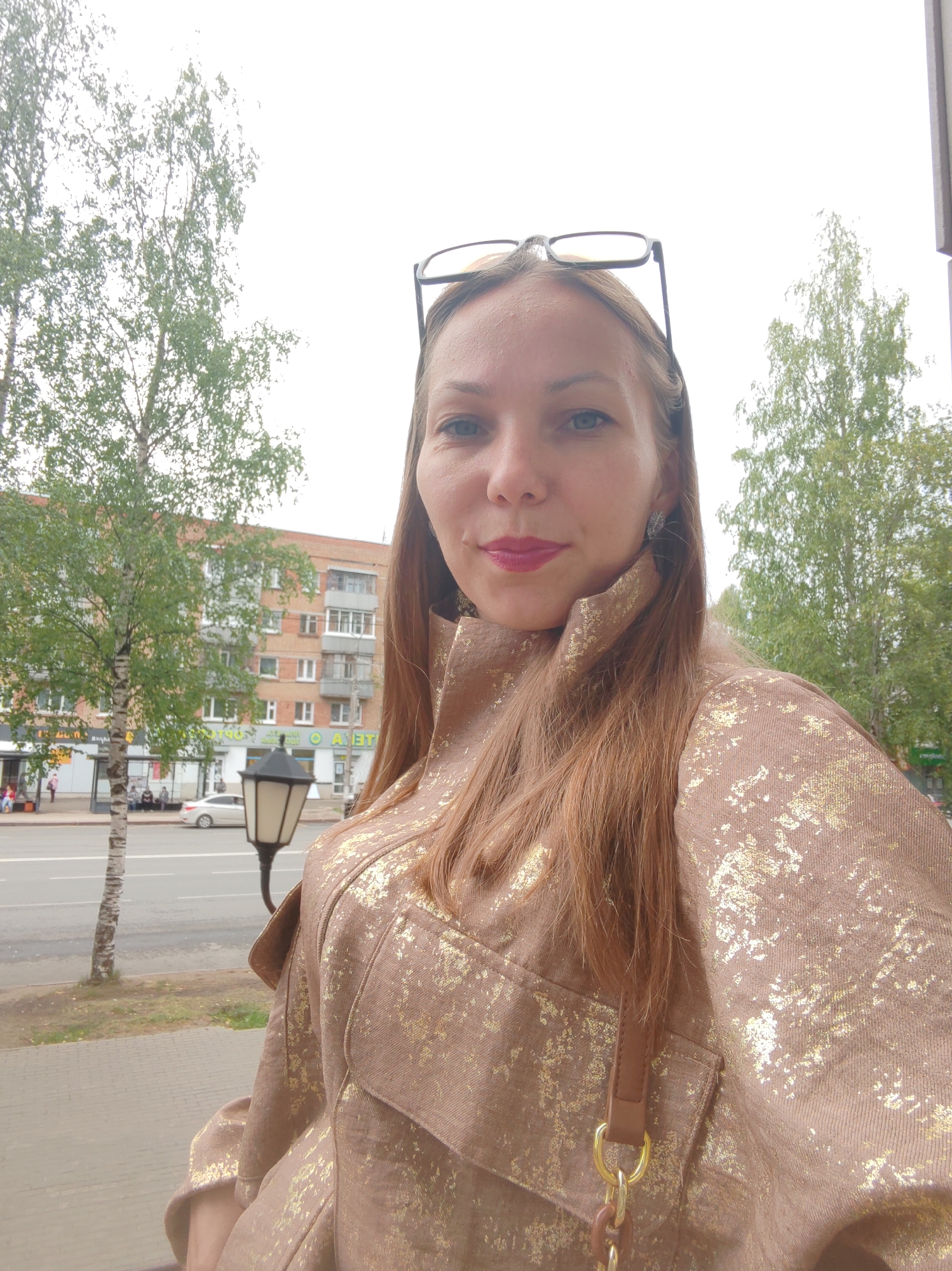 In a medium shot, a woman with long, auburn hair and glasses perched on her head poses for a selfie outdoors. She is wearing a brown jacket with a textured gold pattern and a brown crossbody bag with a gold chain strap. Her makeup includes pink lipstick.
The background suggests an urban environment. To the left, a thin birch tree stands on a paved sidewalk next to a street with a white car driving on it. A street lamp with a black base and a white lantern is visible between the tree and the woman. Further back, a multi-story brick apartment building with several windows and balconies is apparent. The sky is overcast, indicating a cloudy day. The scene is likely a street in Syktyvkar, Russia, given the context of the image.