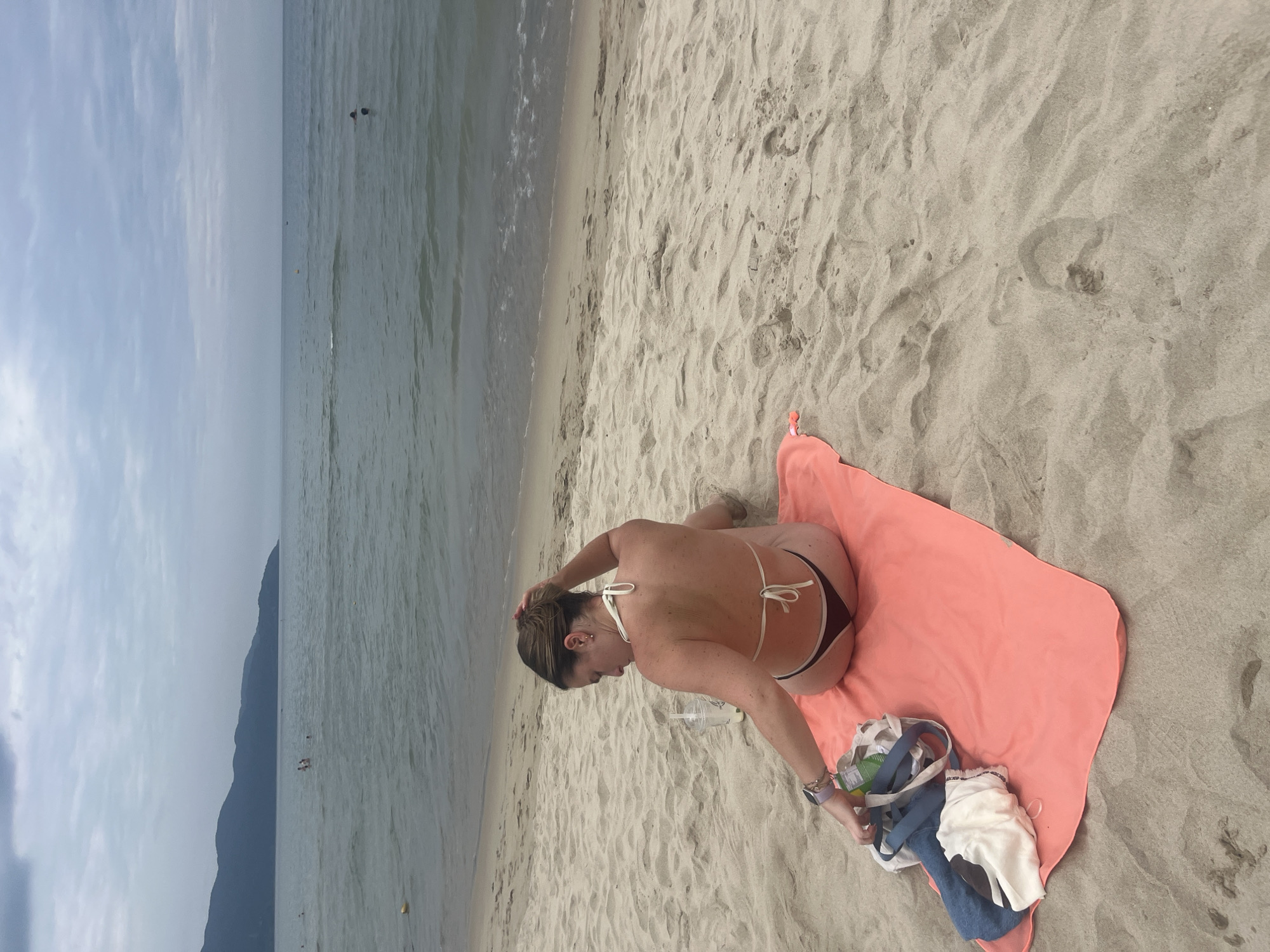 A woman sits on a coral-colored towel on a sandy beach, facing away from the camera. She is wearing a white bikini top and dark bikini bottoms. Her hair is pulled back in a bun, and she is touching it with her right hand. A clear plastic cup with a straw is to her left, and a blue and white tote bag lies beside her. The ocean is in front of her, with a distant mountain range on the horizon. The sky is overcast. This scene is likely on My Khe Beach in the city of Đà Nẵng, Vietnam, a popular tourist destination known for its long stretches of sandy shoreline and views of the Son Tra Peninsula.