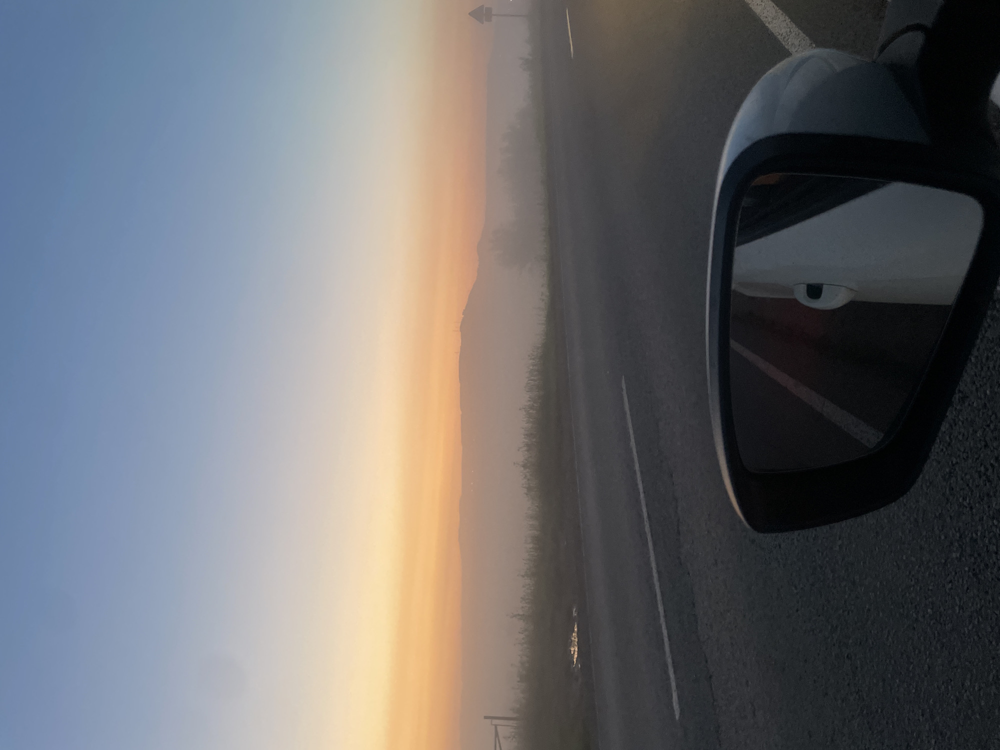 The image depicts a car's side mirror reflecting the interior of the vehicle, including a white door handle and a portion of the car's interior. Outside the car, a road stretches towards a hazy landscape, likely in the city of Uba, Russia, as indicated by the provided context. The scene is bathed in the soft light of dawn or dusk, with a gradient of colors in the sky transitioning from a pale orange near the horizon to a soft blue overhead. A low-lying mist obscures the distant hills and terrain. The road itself is paved and marked with white lines. A few scattered patches of vegetation line the side of the road.