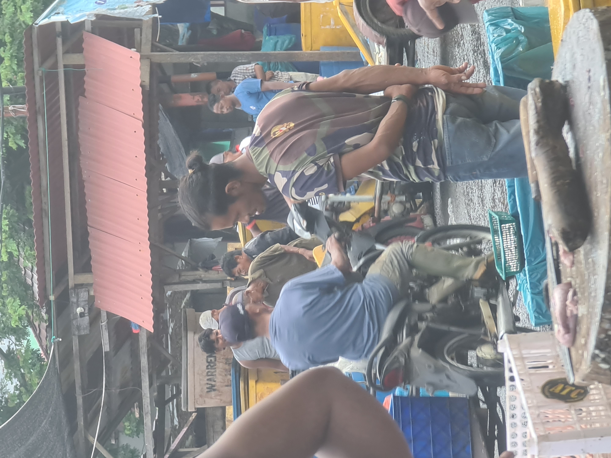 In Lhokseumawe, Indonesia, a bustling market scene unfolds. In the foreground, a wooden chopping block displays raw meat and butchering tools, suggesting a food preparation area. A man, partially obscured by the frame, sits on a motorcycle, facing away from the viewer and wearing a blue t-shirt and khaki pants. His hands rest on the handlebars, and his body is positioned as if about to move.
Behind him, a man in a camouflage t-shirt and blue jeans stands with his hands clasped behind his back, his gaze directed towards the right side of the frame. Several other individuals are present in the mid-ground, some appearing to be engaged in conversation or work activities. Structures with corrugated metal roofs and wooden frames form the backdrop, typical of open-air market environments. Yellow and blue crates are visible, further indicating a commercial setting. The ground is wet and appears to be asphalt or packed dirt, common in outdoor market spaces. The overall atmosphere suggests an active and working marketplace.