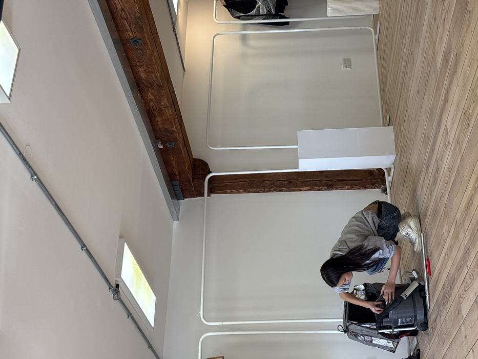 The image portrays an interior scene, likely a commercial studio or retail space, situated in the City of London, United Kingdom. A female individual with long, dark hair, dressed in a grey hooded sweatshirt, distressed blue jeans, and light-colored high-top sneakers, is crouched on a light-toned wooden plank floor. She is actively manipulating components of a black and grey canister-style vacuum cleaner positioned in front of her. A silver-colored metal vacuum extension wand and a small red hand tool are laid on the floor adjacent to the vacuum. To the left, a black bag with a grey and white pattern is partially visible. The background consists of plain white walls. On the right side, two minimalist white clothing racks are present; one supports a dark garment featuring a light geometric pattern. A white rectangular display pedestal stands upright between the racks. The space incorporates rustic architectural elements, including a large, dark brown rough-hewn wooden support beam traversing the upper middle section, consistent with an older renovated building. The white ceiling features exposed grey metal electrical conduit and rectangular light fixtures. A white double electrical outlet is visible on the lower right wall.