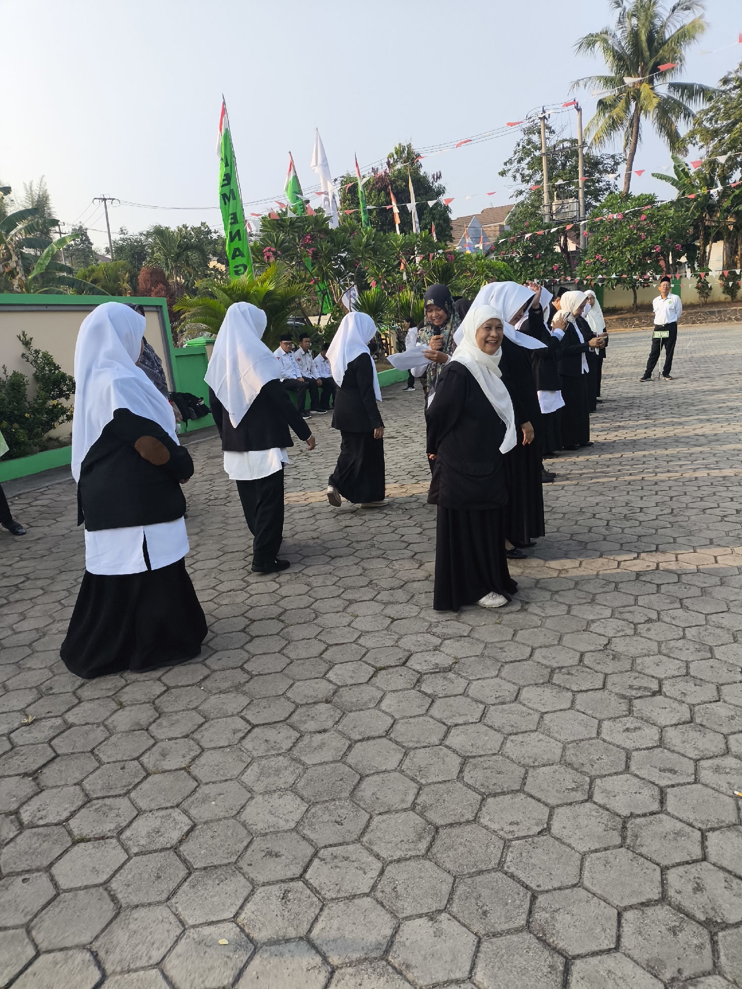 In the city of Cilegon, Indonesia, a group of individuals, predominantly women in black and white attire with white hijabs, are gathered outdoors on a paved hexagonal surface. Several individuals are facing the camera, with one woman in the foreground smiling broadly. Behind them, a man in a white shirt and black pants, possibly a staff member or official, stands with a sign. Trees and flags, including a prominent green flag with white lettering, are visible in the background, suggesting a festive or ceremonial occasion. The setting appears to be a school or public area, with buildings and power lines also present.