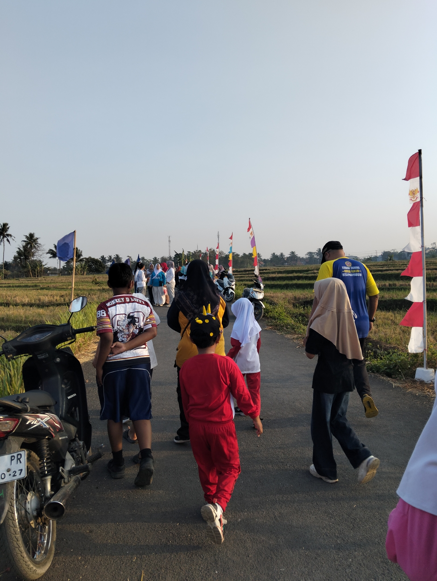 The image shows a group of people walking on a road in a rural area, possibly a rice paddy field, with palm trees and distant structures visible. The sky is clear and light blue. In the foreground, a black motorcycle is parked on the left side of the road. Several individuals are walking away from the camera.
The people are of various ages and are dressed in casual clothing. One person in the foreground is wearing a red outfit with a yellow hat. Another person, a child, is wearing a white shirt with a graphic print and blue shorts. There are also adults in the group, some wearing headscarves.
Along the right side of the road, there are several flags on poles, some red and white, and others with different colored designs. The road appears to be paved. The environment suggests a warm, possibly sunny day.