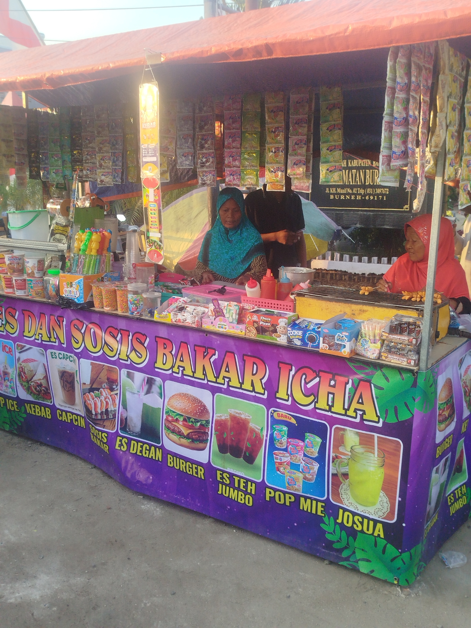 A street food stall is set up under an orange tarp. The stall is selling various snacks and drinks. In the foreground, the stall's banner advertises "IES DAN SOSIS BAKAR ICH A" which translates to "Ice and Grilled Sausage ICHA". Various food items are displayed, including kebabs, sausages, burgers, and drinks like iced tea and what appears to be juice. Packaged snacks such as cookies, candies, and instant noodles are also visible. Two women are attending the stall. One woman, wearing a blue headscarf, is standing behind the counter. Another woman, wearing a red headscarf, is actively grilling sausages on a small grill. In the background, a sign indicates the location: "AH KABUPATEN BURN EH, BURN ER-69171", suggesting the stall is in Burneh, Indonesia. The environment appears to be outdoors, with sunlight illuminating the scene.