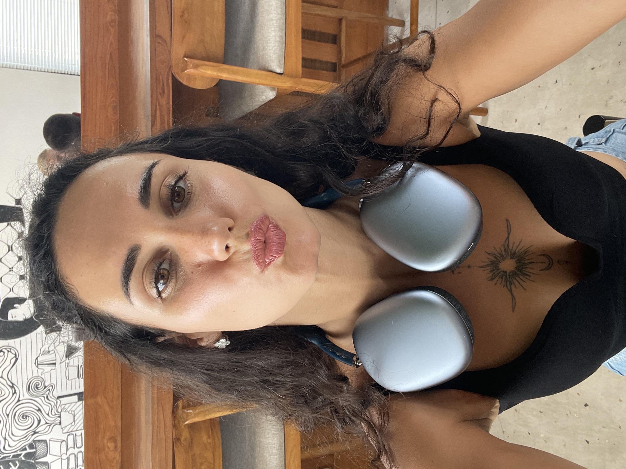 A woman is taking a selfie indoors. She is wearing a black top and has dark, wavy hair. Large, silver headphones are around her neck. A tattoo of a sun and moon is visible on her chest. She is puckering her lips for the selfie. Behind her, there are wooden chairs and tables, suggesting a cafe or restaurant setting. The background also features a mural with a black and white illustration.