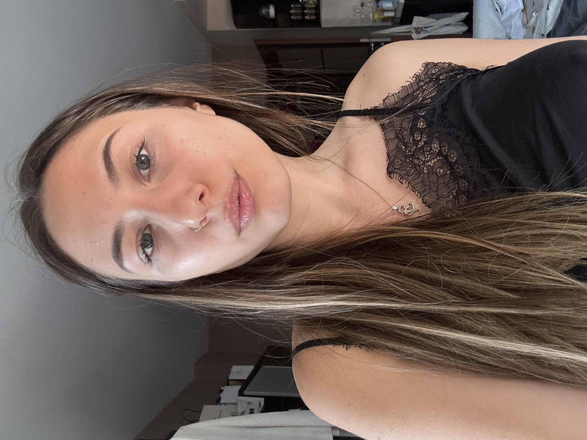 A young woman with long, brown, highlighted hair is pictured in a close-up, selfie-style shot. She is wearing a black lace-trimmed camisole and a silver necklace with a heart-shaped pendant. She has a small nose ring. The background appears to be an indoor setting, possibly a home, with a grey ceiling and a blurred background that includes what looks like clothing hanging on a rack, a television, shelves with various items, and a doorway. The image is not specific to Veliko Tarnovo, Bulgaria, as there are no identifiable landmarks or contextual clues to place it in that location.