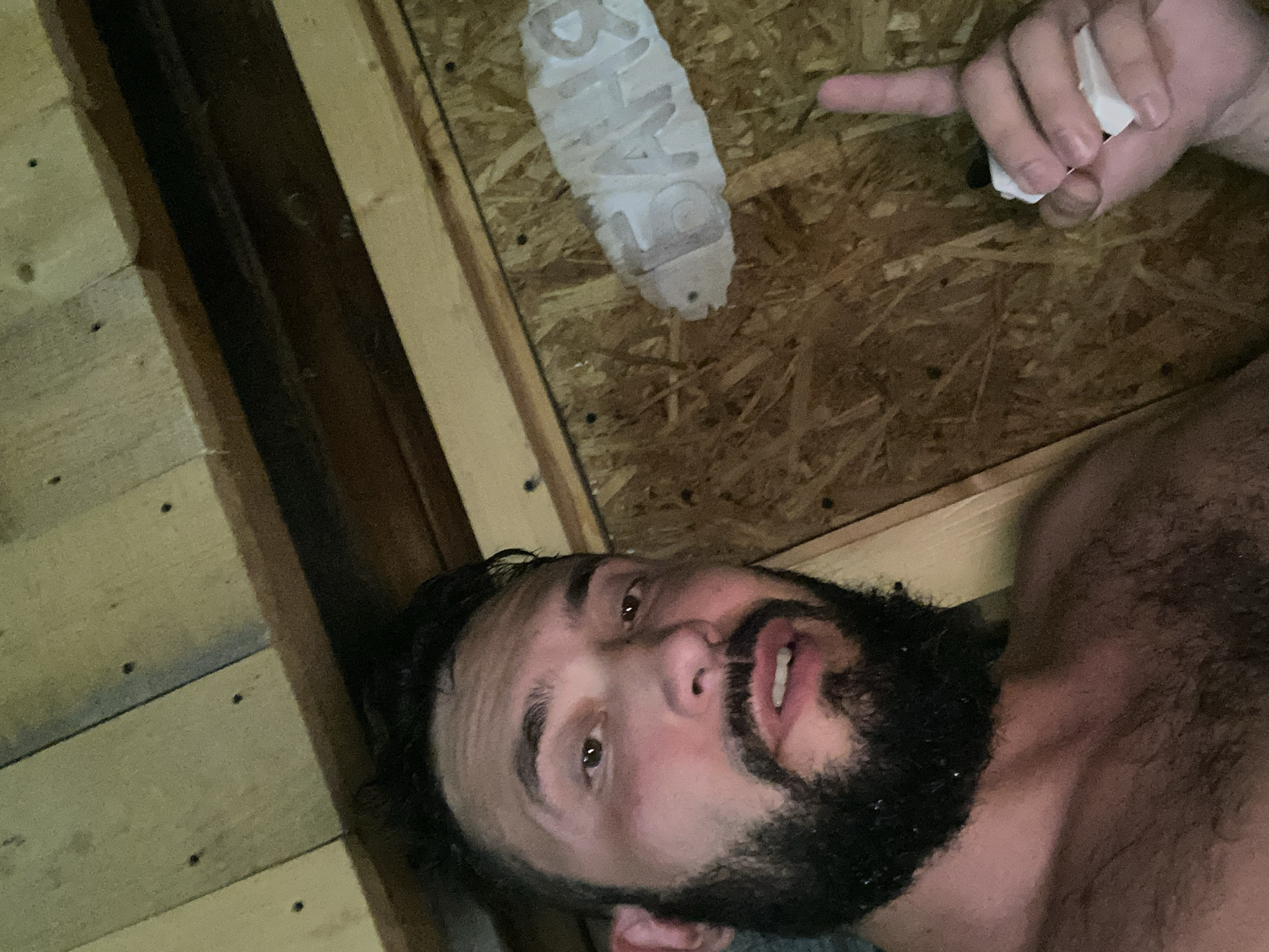 A man with dark hair and a beard is in a wooden room. He is facing the camera and smiling. The room appears to be a sauna, with wooden walls and a ceiling made of planks. There is a sign on the wall that reads "БАЊЯ," which is Russian for "bathhouse." The man is wearing a towel around his waist. He is holding a piece of paper in his right hand and pointing up with his index finger.