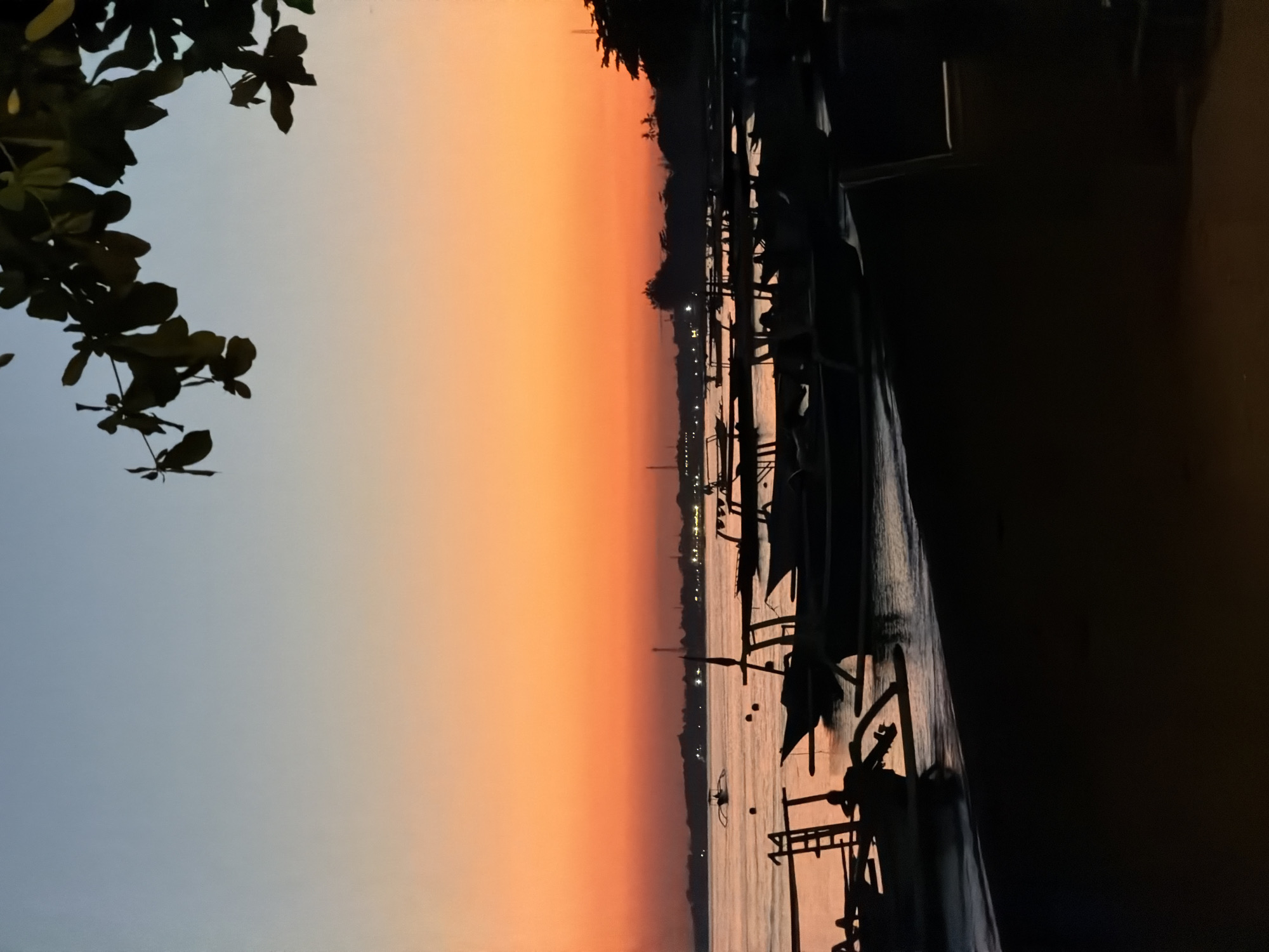 A silhouette of traditional Balinese fishing boats lines the shore of Buleleng, Indonesia, under a sky painted with the warm hues of a sunset. The water reflects the orange and blue gradients of the sky, creating a serene and tranquil atmosphere. In the foreground, several boats are anchored, their dark shapes contrasting with the soft glow of the horizon. A single, larger boat with an outrigger stands out further out in the calm sea. The shoreline is also silhouetted, with a hint of foliage from a tree on the right side of the frame adding a natural element to the scene. The overall image captures the quiet beauty of a coastal evening in Buleleng.