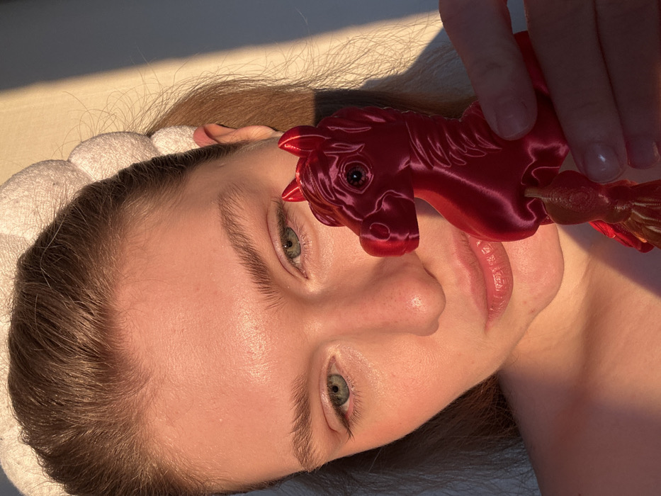 A light-skinned woman lies with her head tilted to the left, wearing a white headband. The sun highlights her face, emphasizing her green eyes, fair skin, and light brown hair. A shiny red horse toy is positioned near her face, held by a hand whose fingers are partly visible. The photo's perspective focuses on the woman's face and the toy, with a soft background.