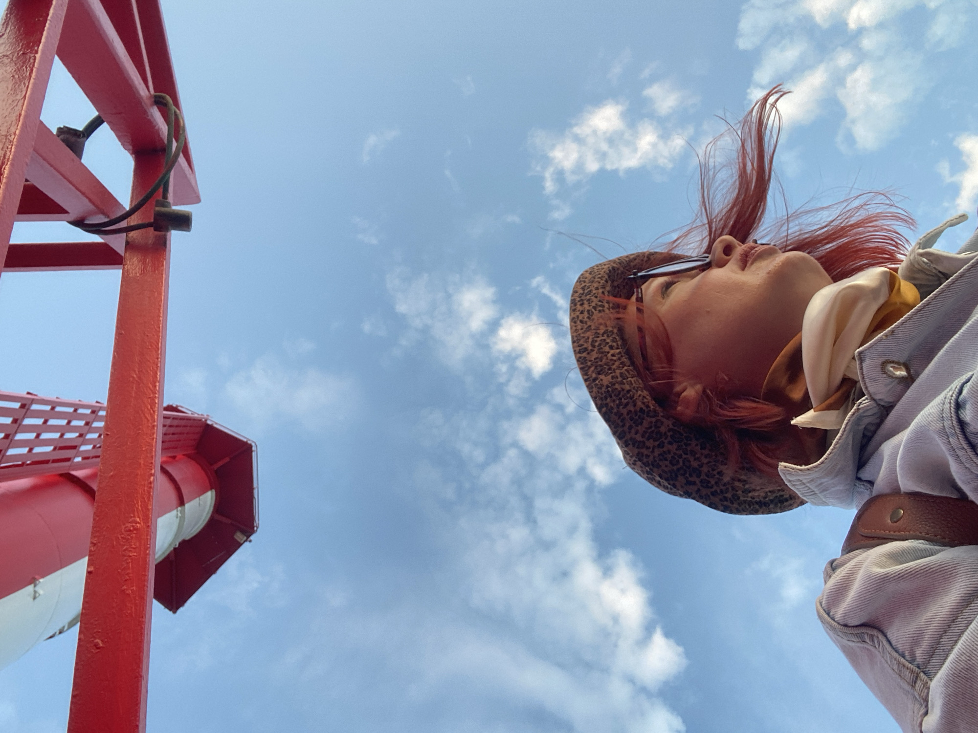The low-angle image shows a person looking up at a red and white lighthouse-like structure against a blue sky with scattered clouds. The person has red hair, wears a leopard print hat and glasses, and a denim jacket with a brown leather belt detail. The lighthouse structure is partially visible on the left side of the frame. The location is Sochi, Russia.