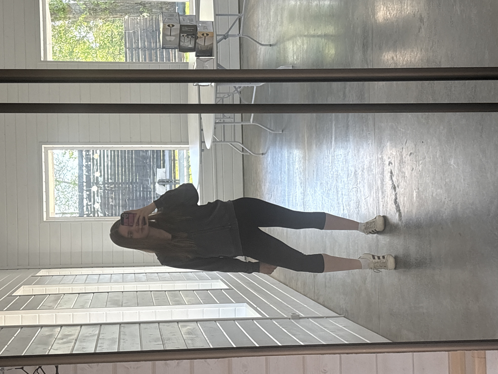 A full-length mirror reflects a woman in workout clothes (jacket and leggings) holding a cell phone. The reflection shows an interior room with white horizontally-paneled walls and a gray concrete floor. Light fixtures are visible in the background through the windows, along with trees. Tables and chairs are also visible. The location is not identifiable as Юг, Russia from the image alone.