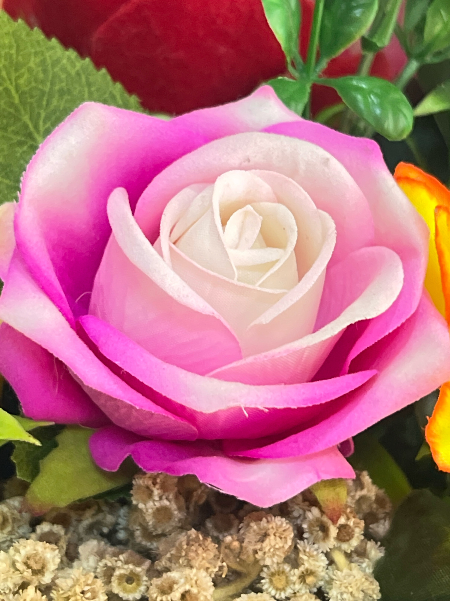 This image is a close-up, low-angle, vertical shot of a pink and white artificial rose. The rose is the primary subject, with its petals appearing soft and layered. The center of the rose is a pale creamy white, transitioning to a soft pink, and then to a vibrant fuchsia pink on the outer petals. The texture of the petals appears to be fabric. Behind the rose, out of focus, are hints of other artificial flowers and greenery, including a blurred red flower at the top and a sliver of an orange and yellow flower on the right. The background is dark and indistinct, suggesting an indoor setting or a floral arrangement. There is no indication of the city of Denpasar, Indonesia, or any people, actions, or interactions.