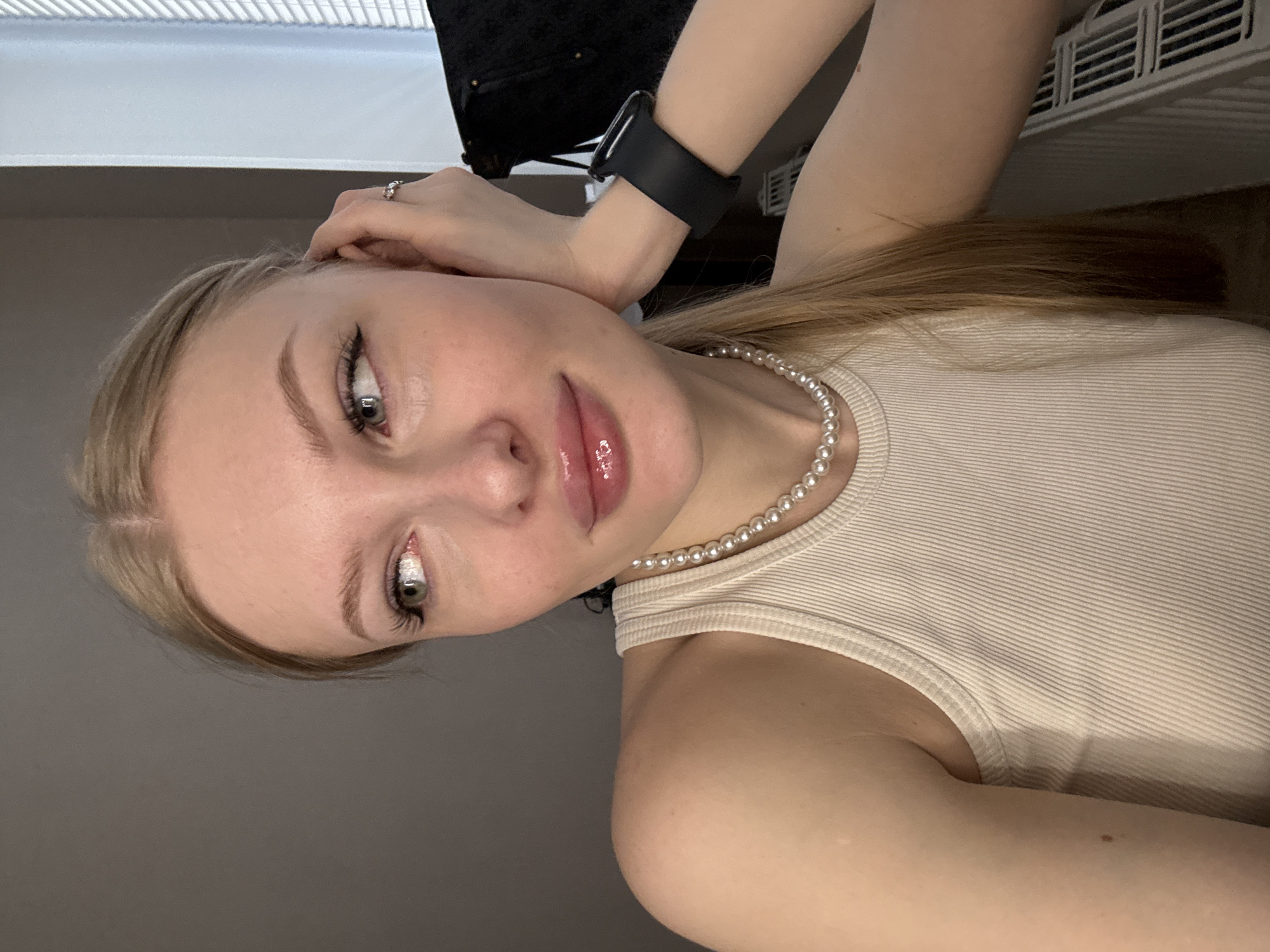 The image depicts a close-up, eye-level, portrait-style shot of a young Caucasian woman with blonde hair, wearing a cream-colored ribbed tank top and a pearl necklace. She is positioned in what appears to be an indoor setting, with a neutral gray wall as the background on the left and a window with blinds on the right. Her right hand is supporting her head, with a ring visible on her ring finger and a black smartwatch on her wrist. Her gaze is directed to her left, away from the camera. The lighting suggests an indoor environment, possibly artificial, with some natural light filtering in from the window. There is no indication of the city of Perm, Russia, within the image.