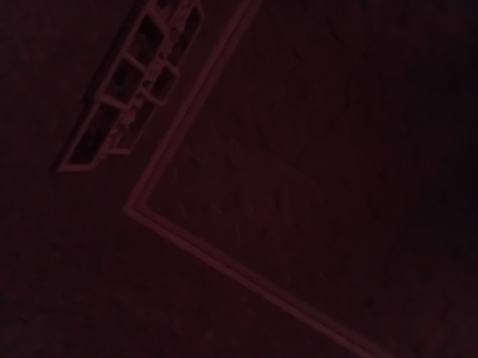 The image shows a dark, possibly interior, scene with a faint red light illuminating some structural elements. In the upper right portion of the image, a segmented, rectangular object with multiple openings is visible, suggesting it could be part of a ceiling fixture or an electrical panel. A line of this red light also forms a corner or outline on a textured, dark surface in the lower and central parts of the image, possibly indicating a border or a guide. The overall impression is one of low visibility and an unfinished or industrial environment. There are no people or animals visible, and no discernable activity. The location in Verkhniachka, Ukraine, is not visually represented in the image itself.