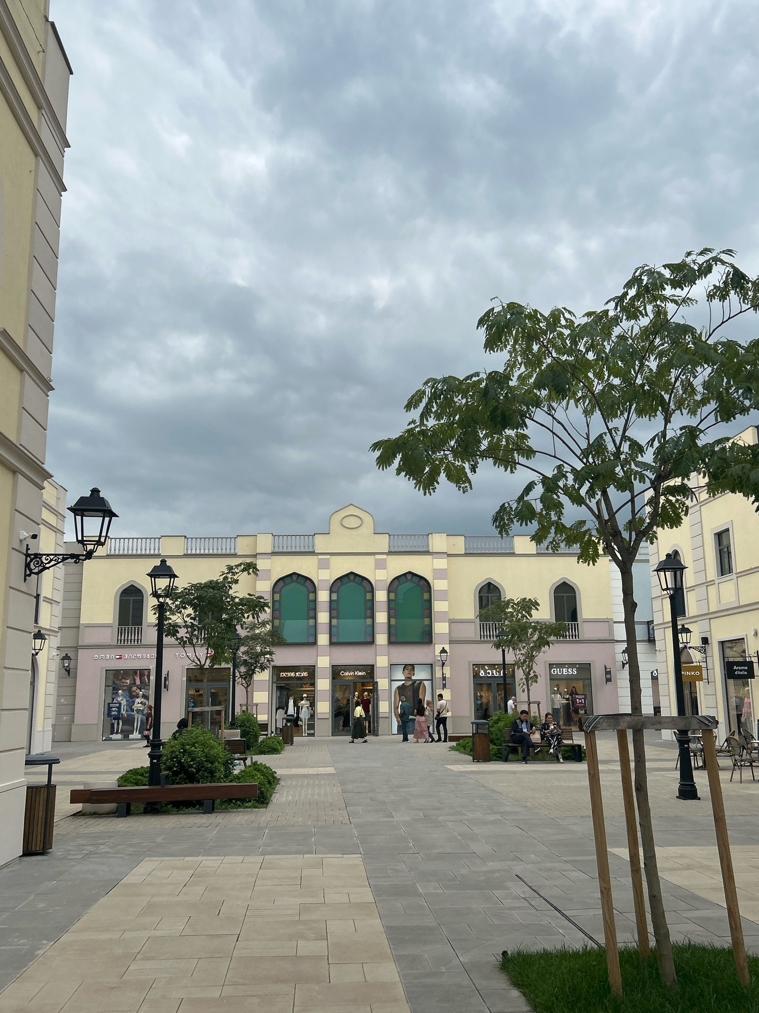 The image shows an outdoor shopping area with a variety of retail stores lining a pedestrian walkway. The architecture of the buildings is characterized by light yellow facades, arched windows, and decorative balustrades. Storefronts visible include Tommy Hilfiger, Guess, and Calvin Klein. People are seen walking, sitting on benches, and browsing in front of the shops. The walkway is paved with light-colored stone tiles, and there are streetlights and trees integrated into the landscape. The sky is overcast and cloudy, suggesting a temperate climate. This scene is located in Gamarjveba, Georgia, likely an outlet mall or a similar commercial development.